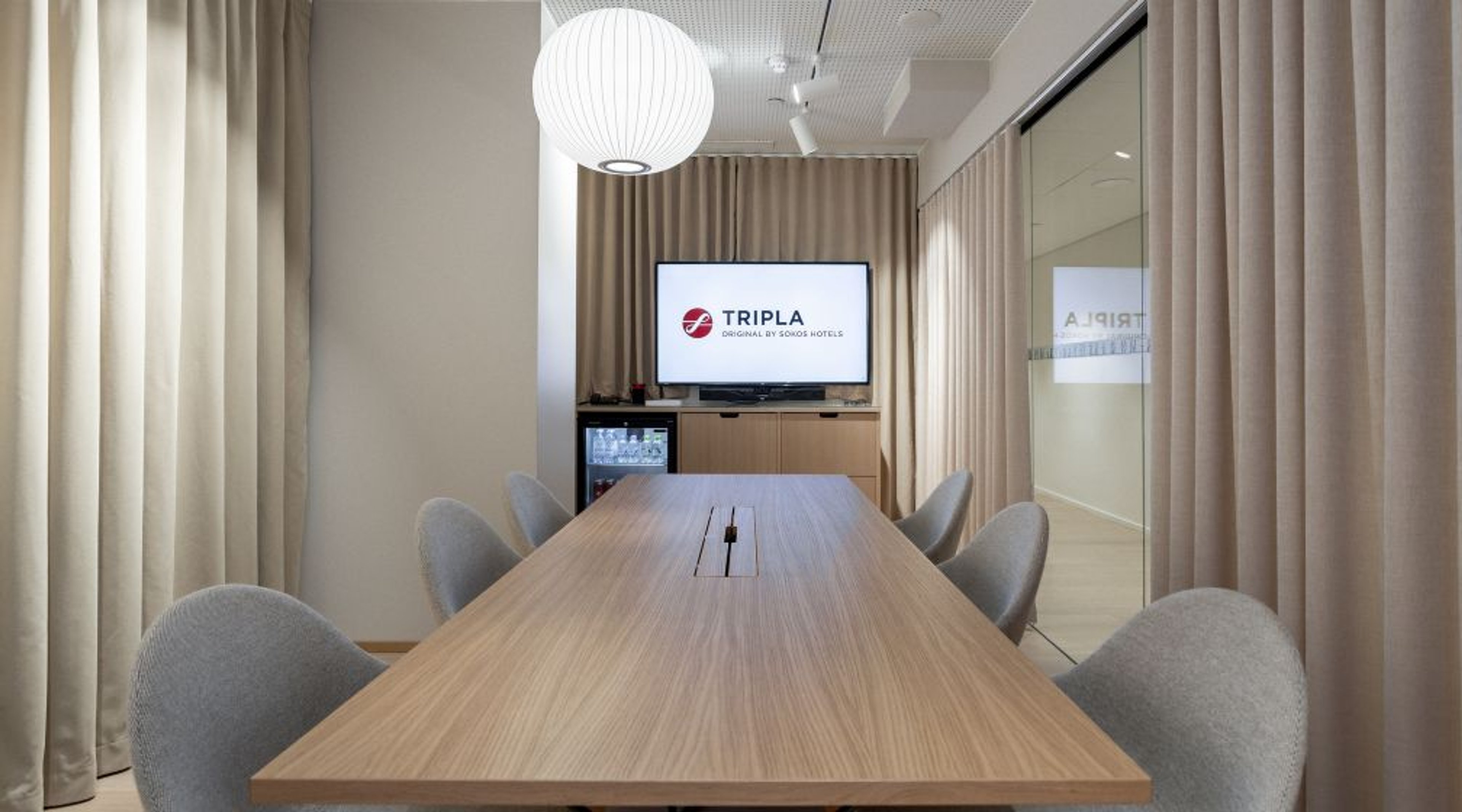 Moderni neuvotteluhuone, jossa puinen pöytä, harmaat tuolit, beige verhot, valkoinen pallomainen kattovalaisin ja tv-näyttö seinällä.