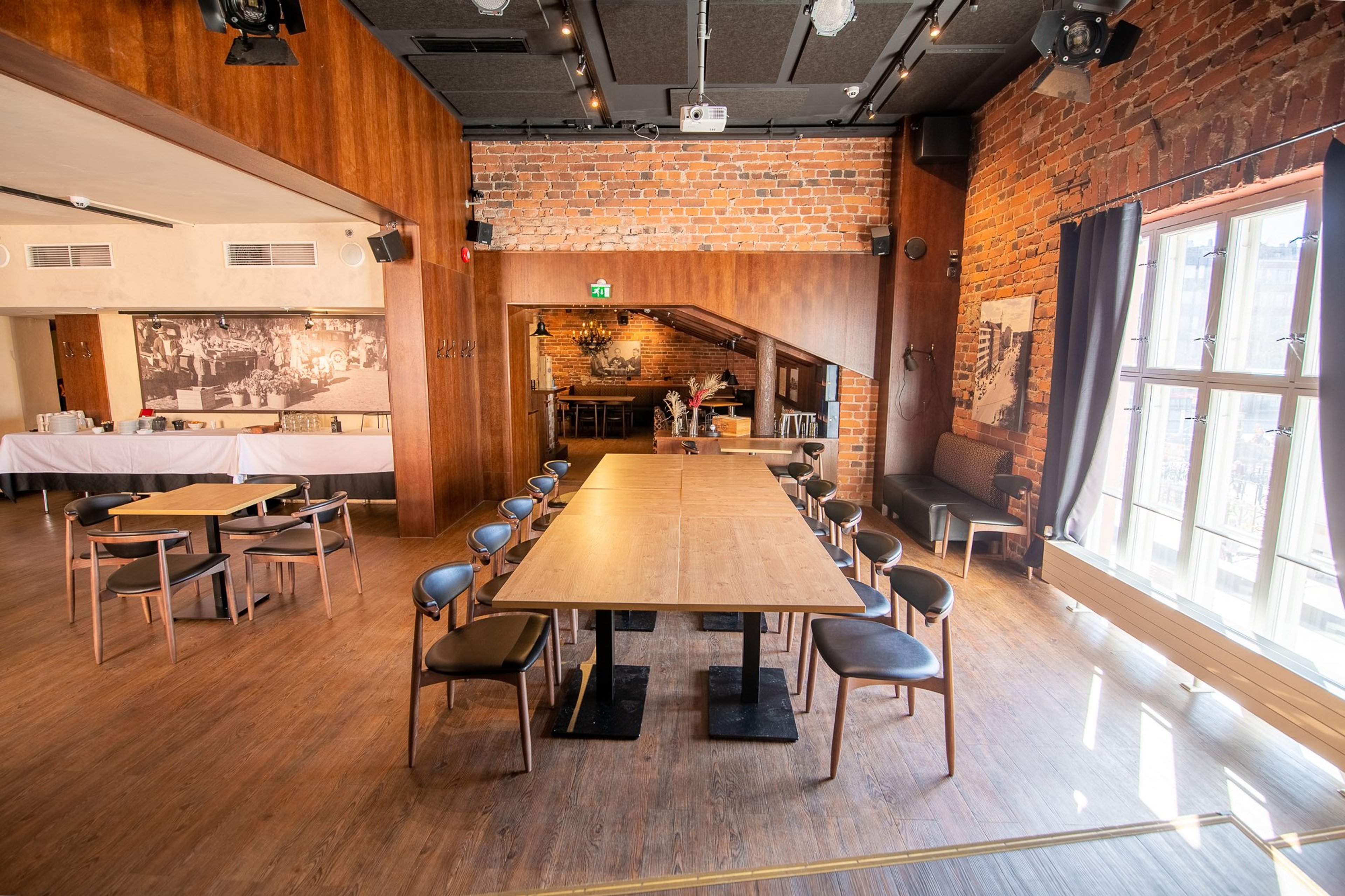 Kokoustila tiilisinillä seinillä, puulattialla ja suurilla ikkunoilla. Pitkä puupöytä tuoleineen ja näkymä viereiseen tilaan.