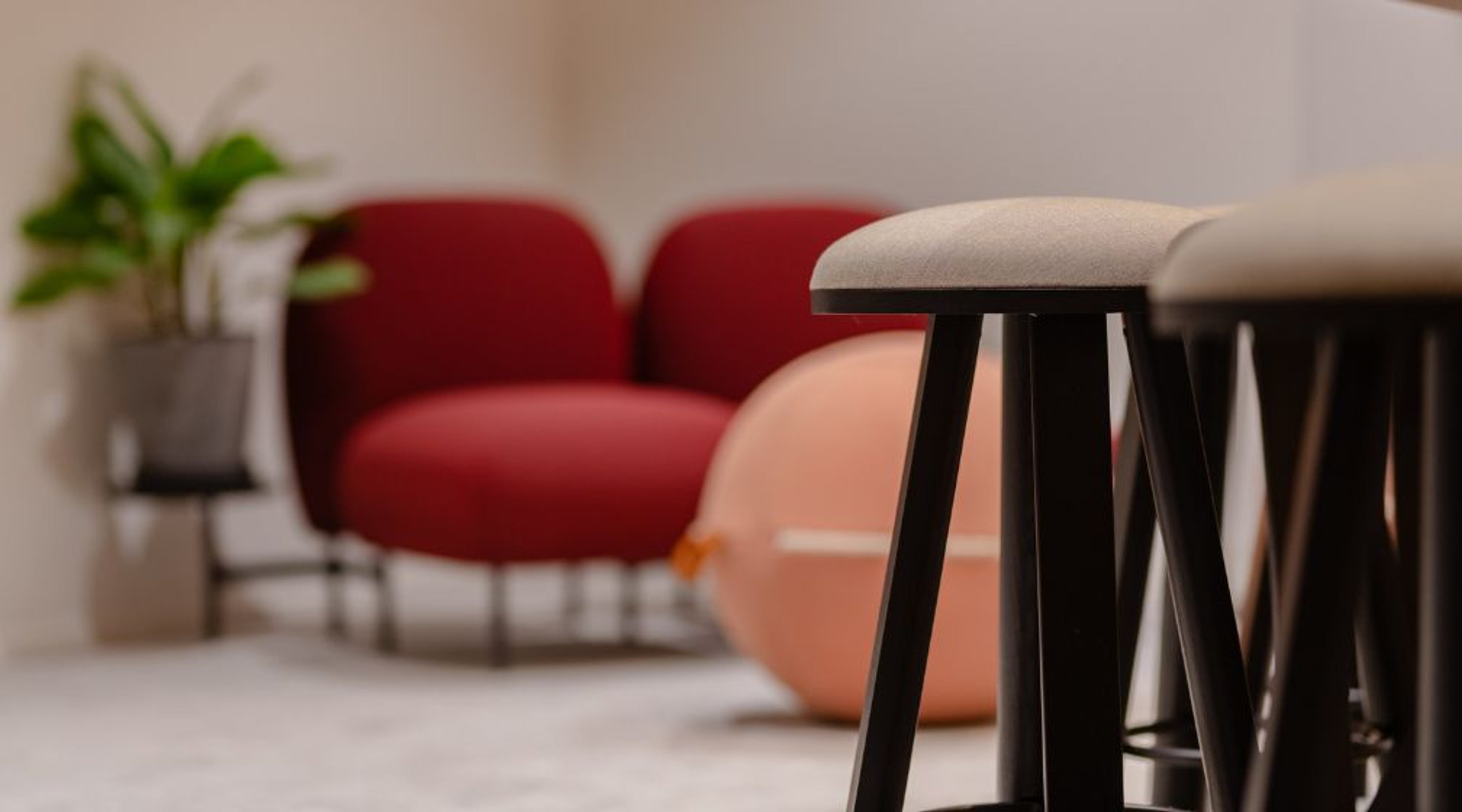 Moderni kokoustila, jossa vaalea jakkara mustilla jaloilla etualalla, taustalla punaisia ja vaaleanpunaisia nojatuoleja sekä viherkasvi.