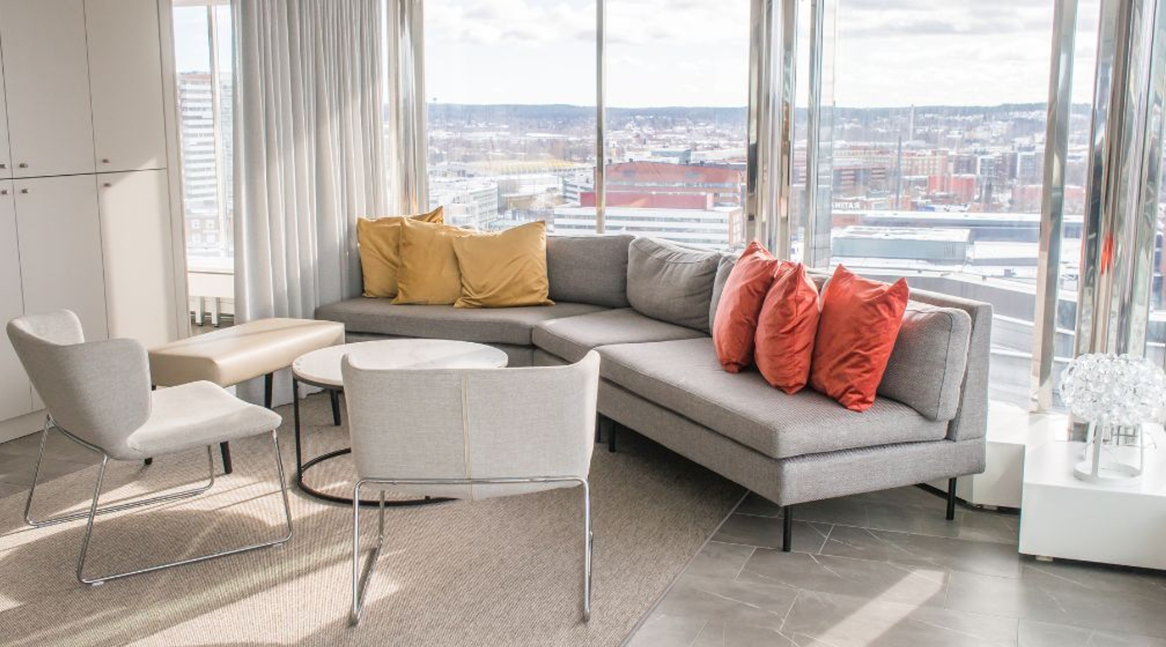 Moderni olohuone, jossa harmaa kulmasohva oransseilla ja keltaisilla tyynyillä. Isot ikkunat tarjoavat kaupunkinäkymän. Huoneessa vaalea lattia ja valkoisia tuoleja.