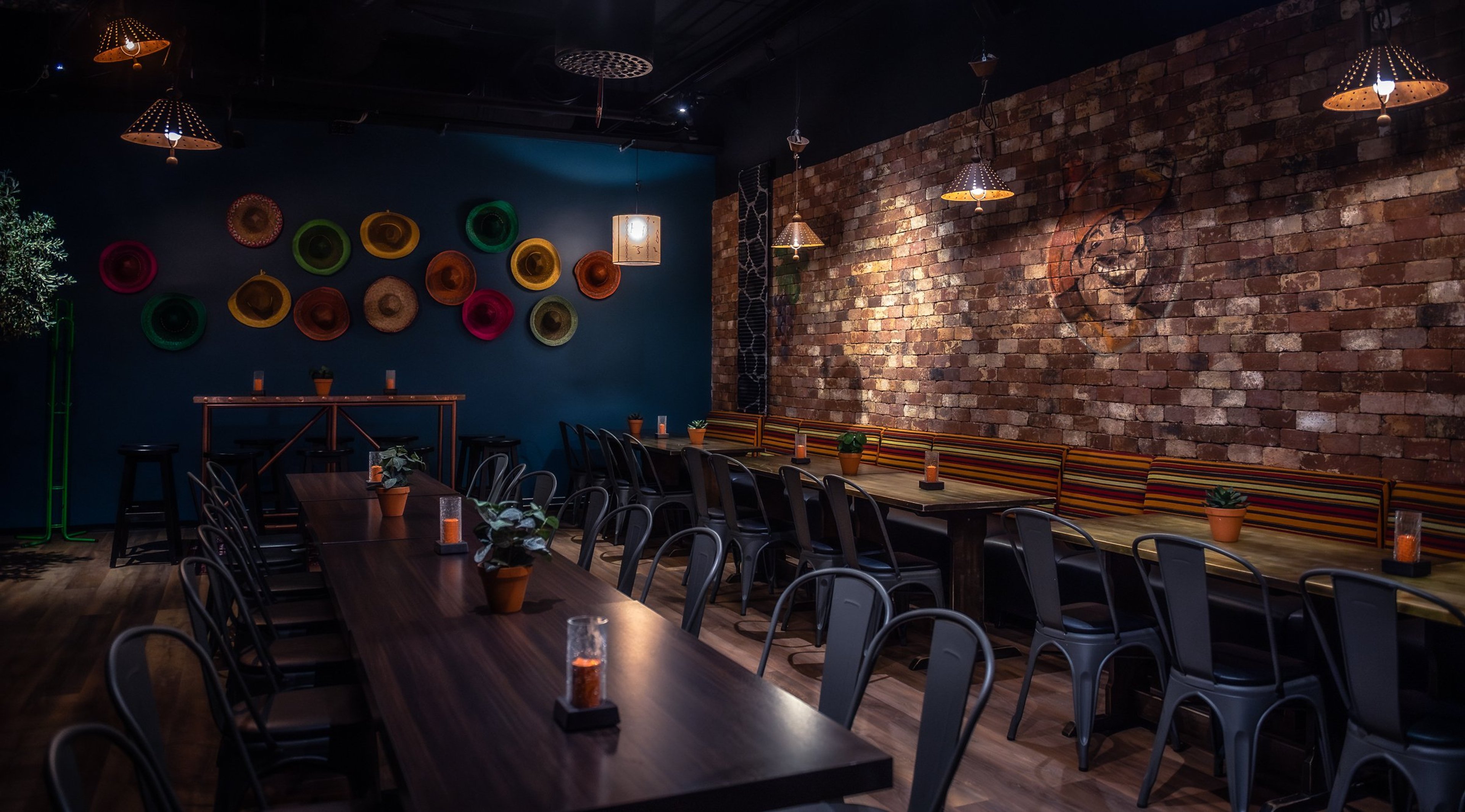 Tunnelmallinen ravintola, jossa tummansininen seinä värikkäillä hatuilla, tiiliseinä ja mustat metallituolit puupöytien ympärillä. Kattovalaisimet ja puulattia.