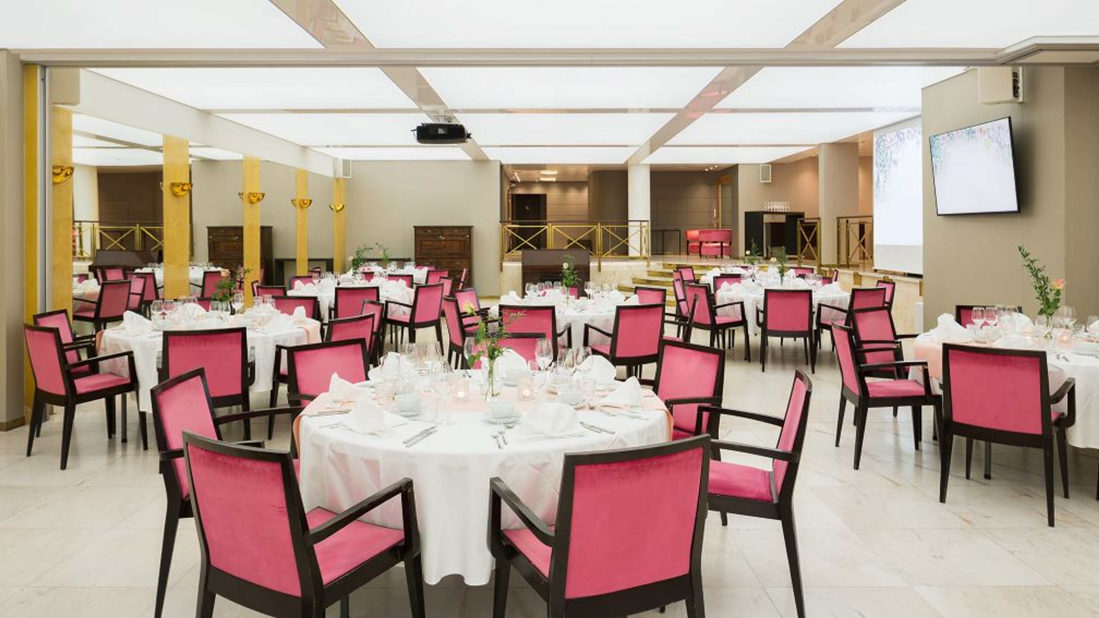 Juhlatila, jossa pyöreitä pöytiä valkoisilla liinoilla ja kirkkaanpunaisia tuoleja. Tilassa tasainen marmorimainen lattia ja kattoprojektori.