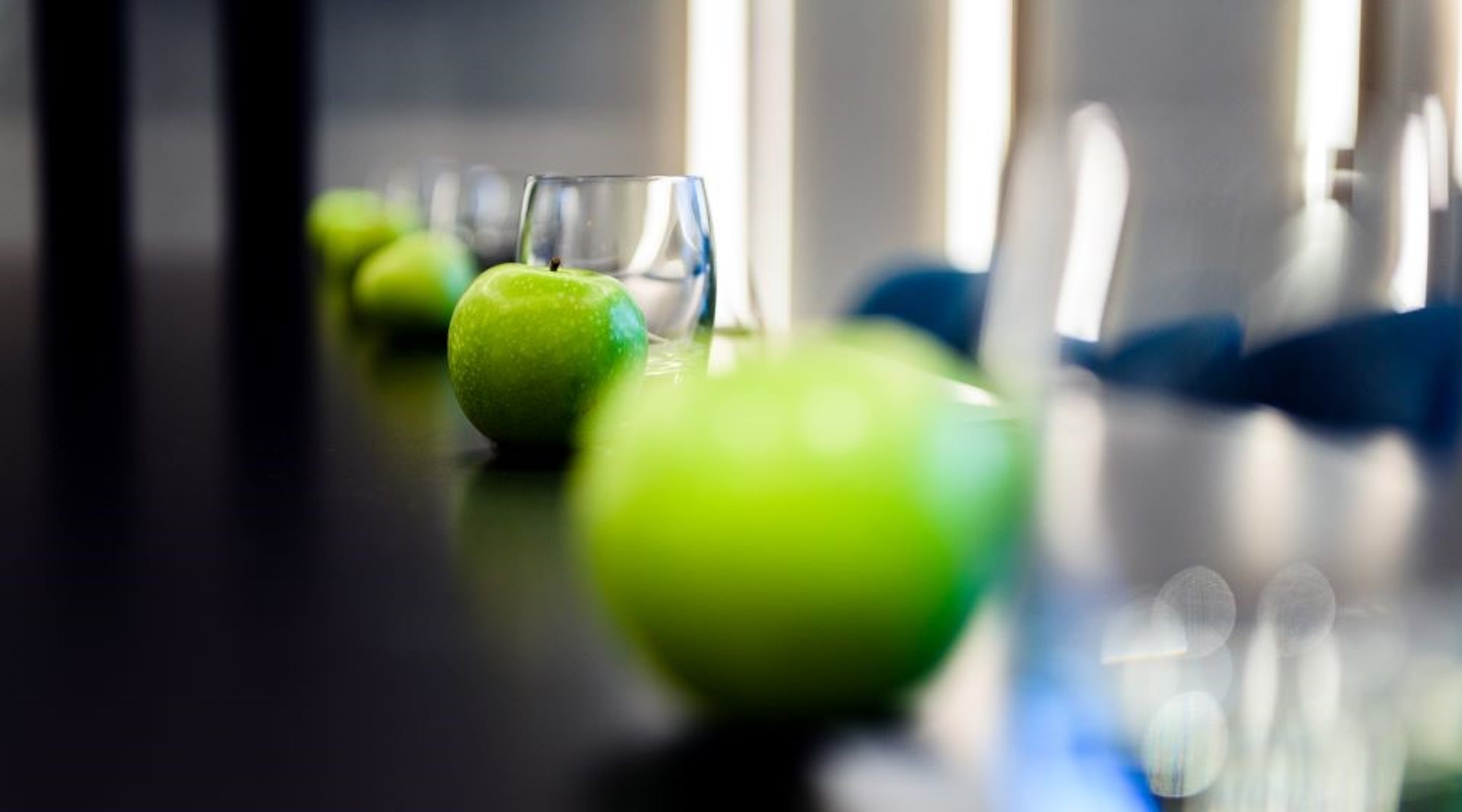 Vihreitä omenoita ja vesikannu rivissä tummalla kokouspöydällä, taustalla näkyy vaaleita tuoleja.