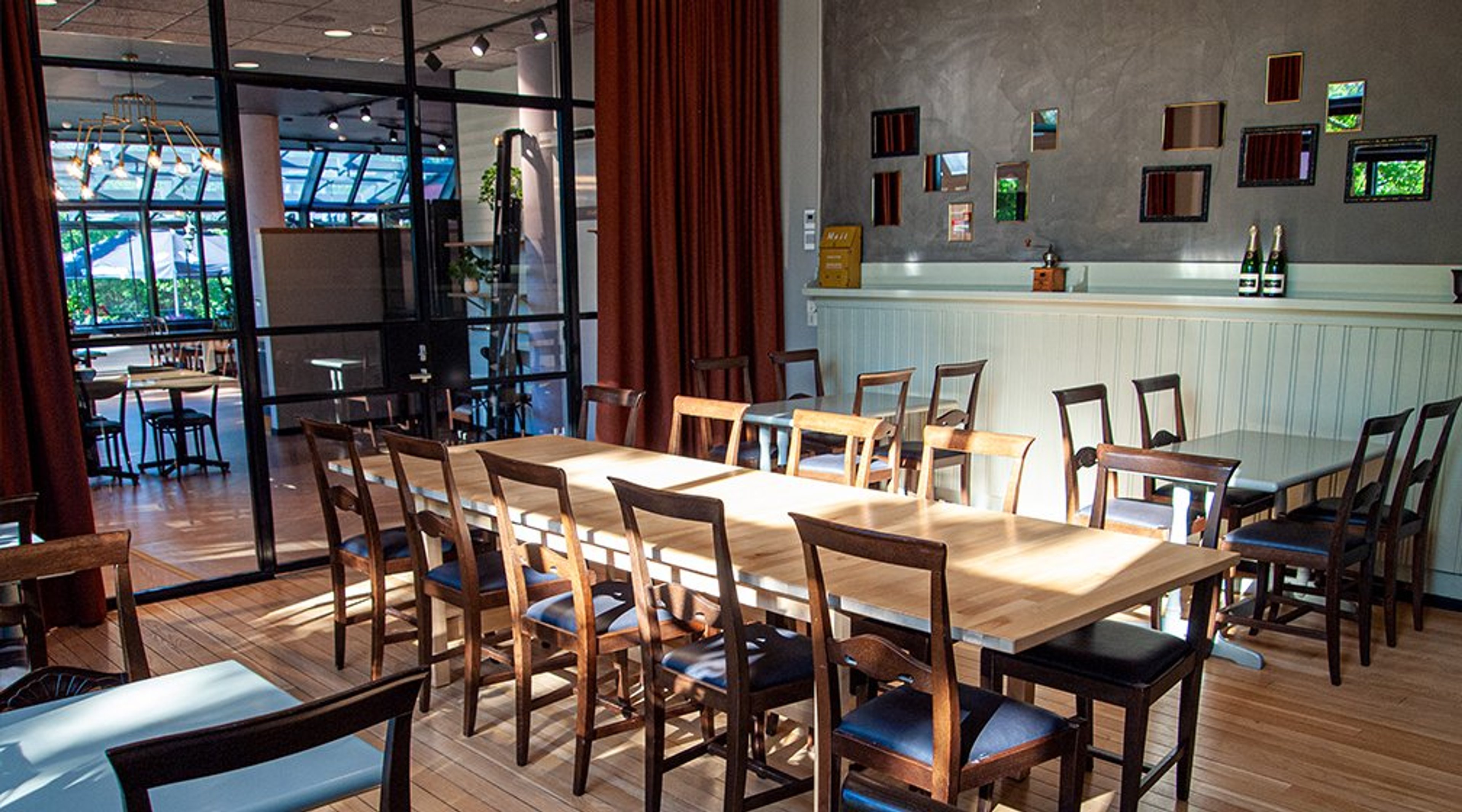 Ravintolan yksityinen tila, jossa pitkä puinen pöytä tuoleineen. Taustalla lasinen väliseinä, punaiset verhot ja koristeellinen harmaa seinä pienillä ikkunoilla. Puulattia ja vaalea baaritiski.