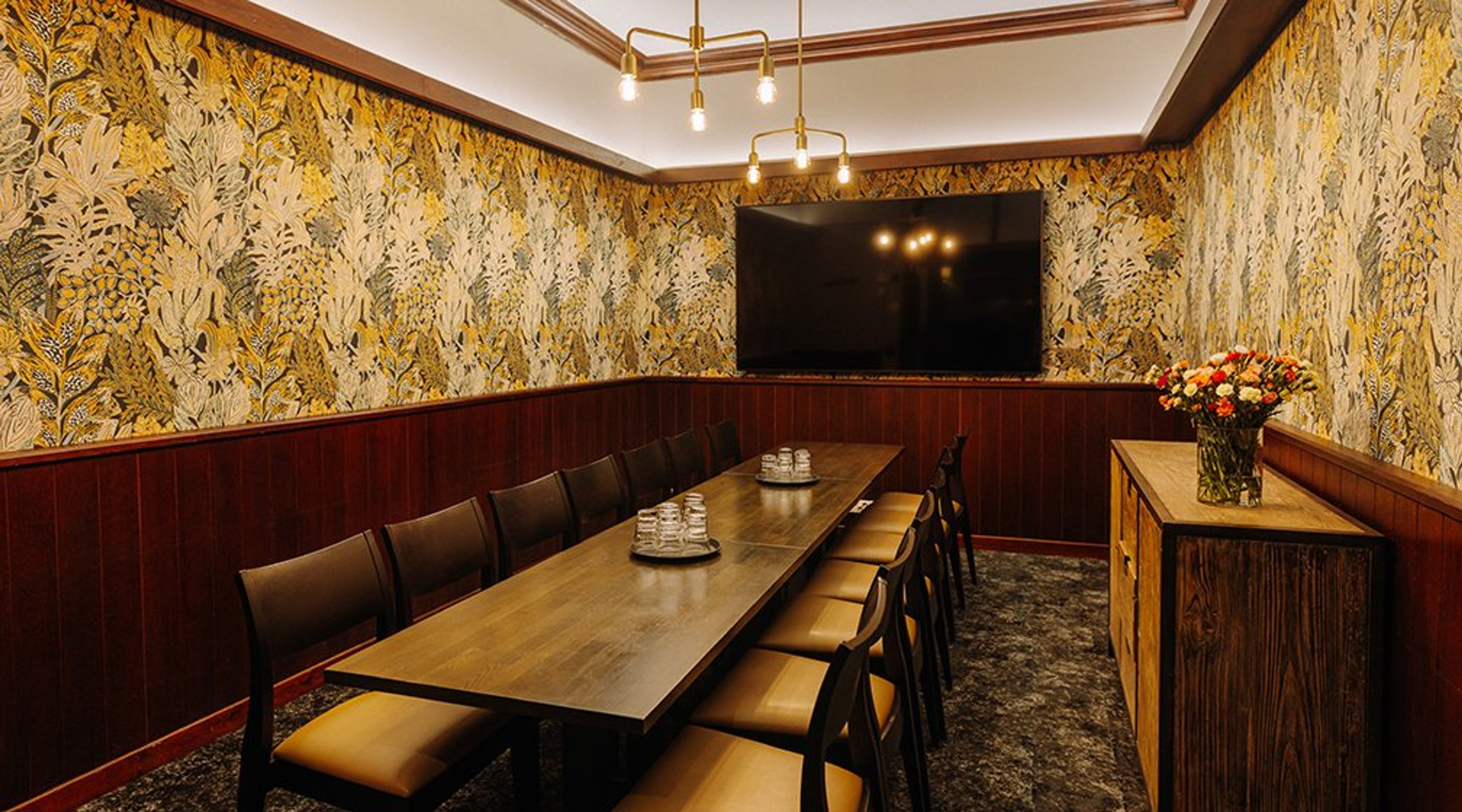 Kokoushuone, jossa pitkä puinen pöytä tuoleineen, keltakukkainen tapetti ja tv-näyttö. Seinällä puupanelointi ja sivupöydällä kukkakimppu.