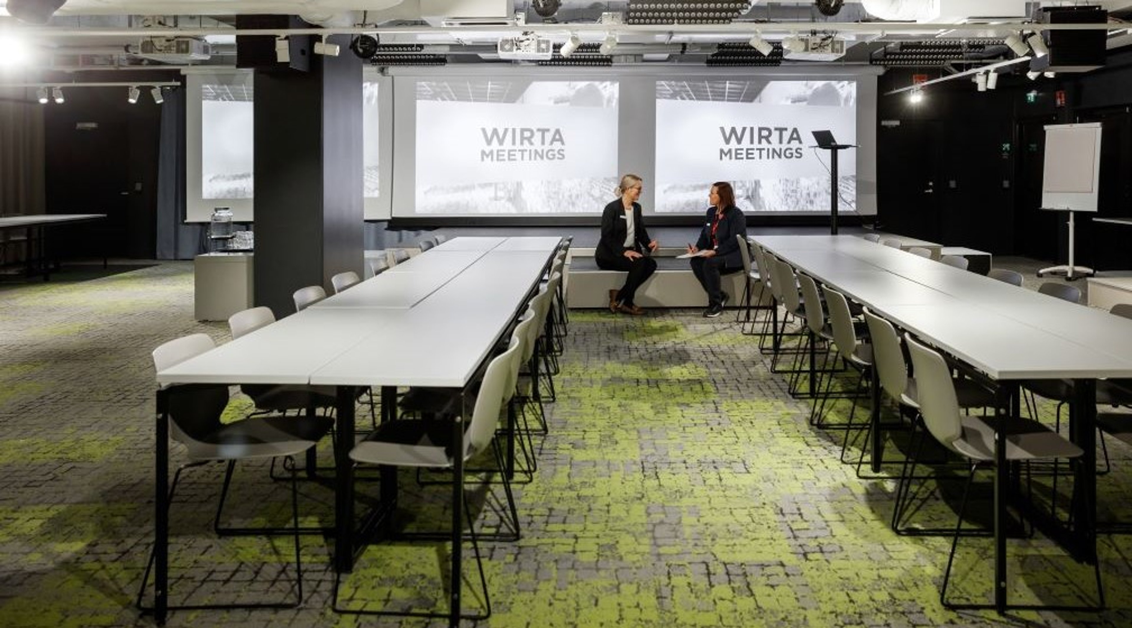 Moderni kokoustila pitkillä pöydillä ja valkoisilla tuoleilla. Taustalla näkyy isot näytöt, kaksi henkilöä keskustelee taustalla. Lattia on värikkäästi kuvioitu.