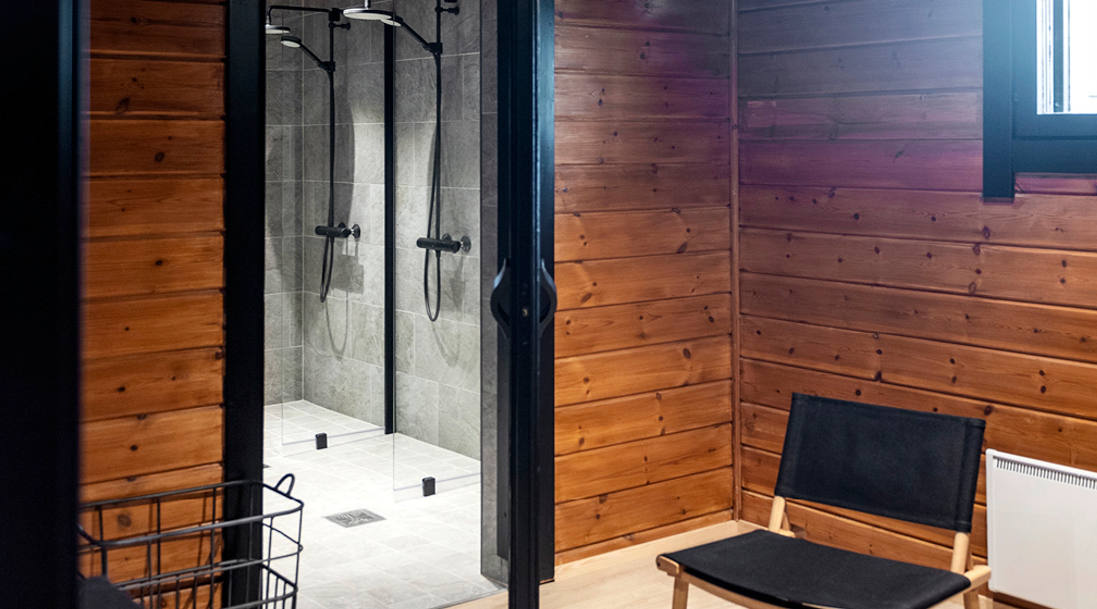 Moderni kylpyhuone puupaneeliseinillä. Lasiovellinen suihkutila kahdella suihkulla ja musta tuoli oikealla.