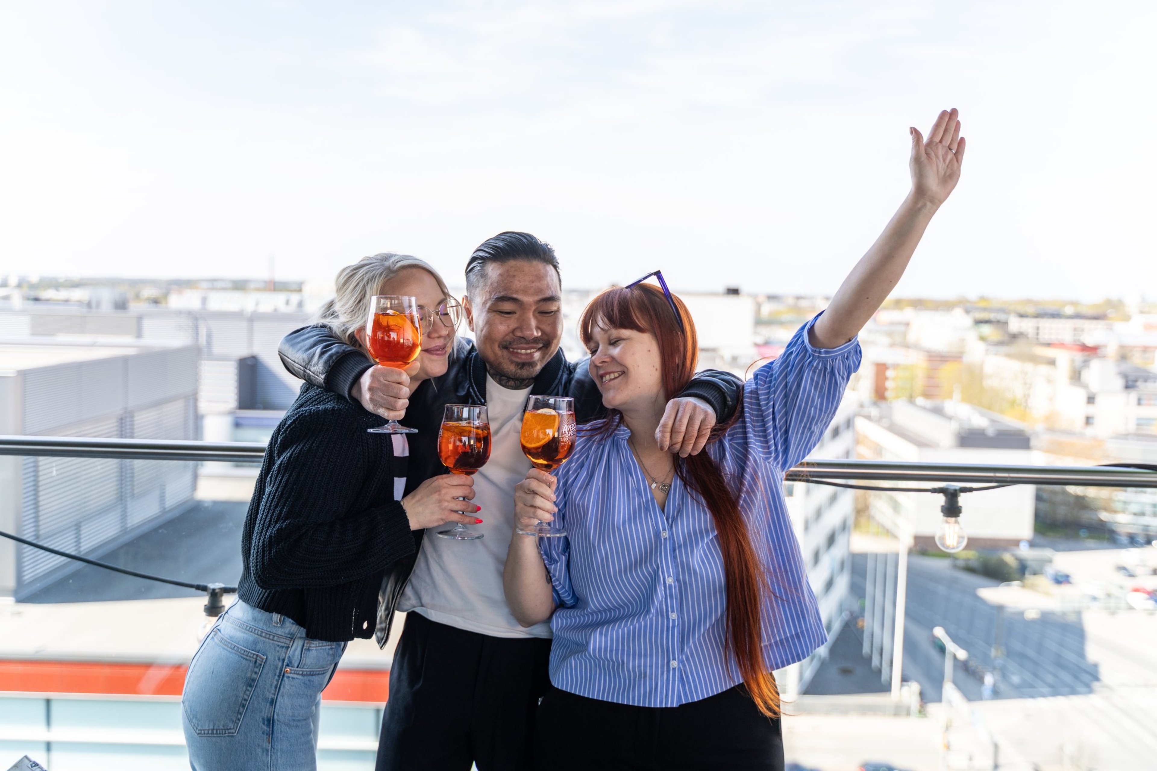 Kolme henkilöä nauttimassa juomia aurinkoisella kattoterassilla, josta avautuu näkymä kaupunkiin.
