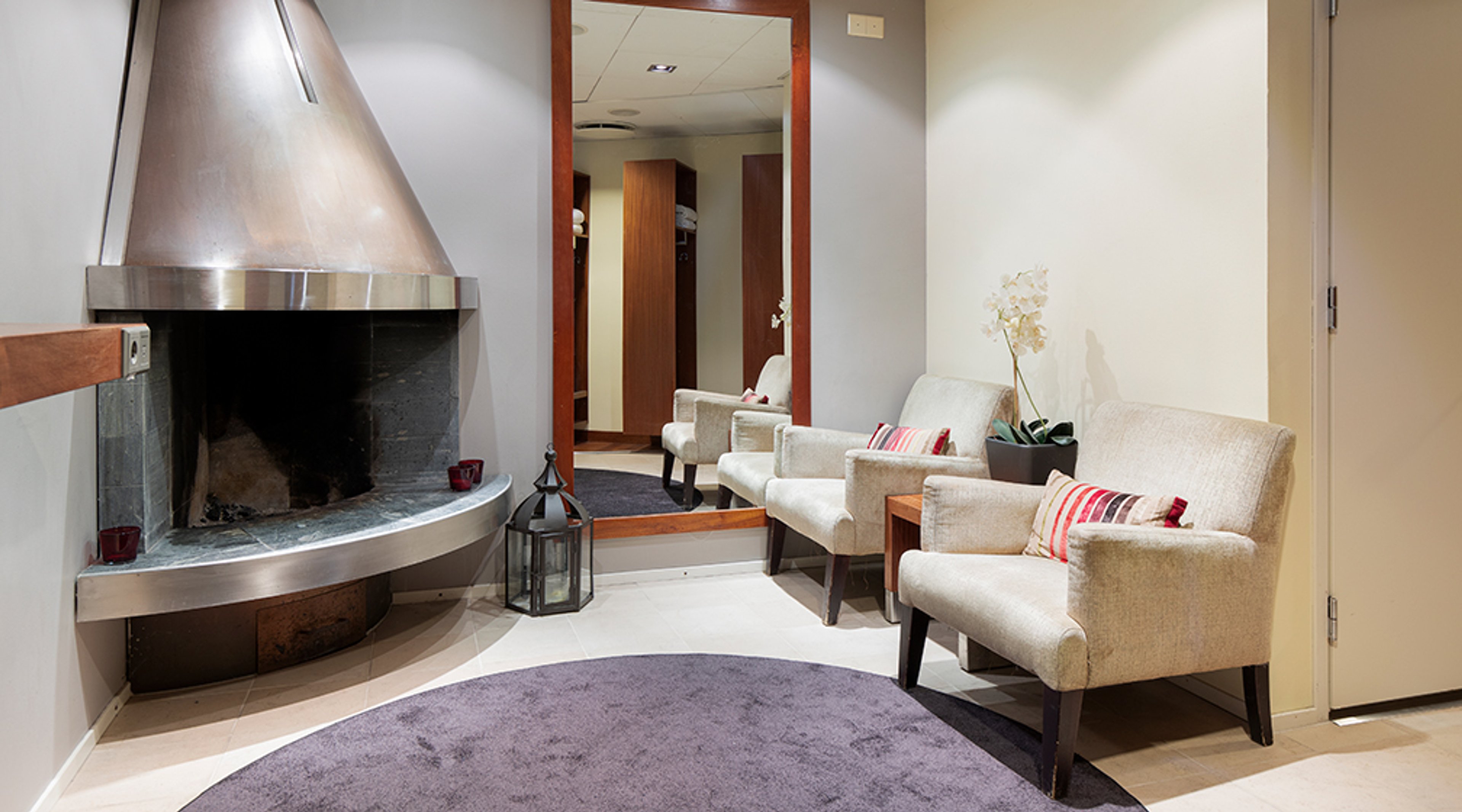 Taukotila, jossa moderni takka, kaksi vaaleaa nojatuolia punaisilla tyynyillä, pyöreä violetti matto ja käytävänäkymä peilissä.