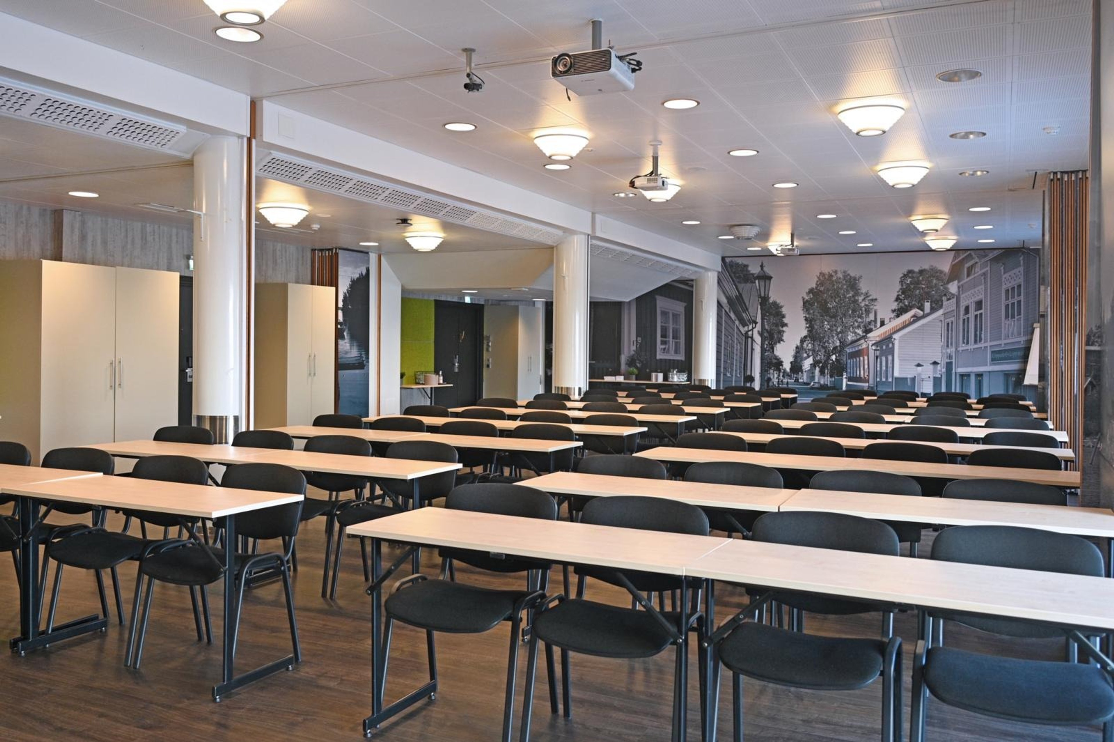 Luokkahuonemainen kokoustila, jossa pitkät pöydät tuoleineen riveittäin, puulattia ja takaseinällä historiallisen kaupunkimaiseman kuva.