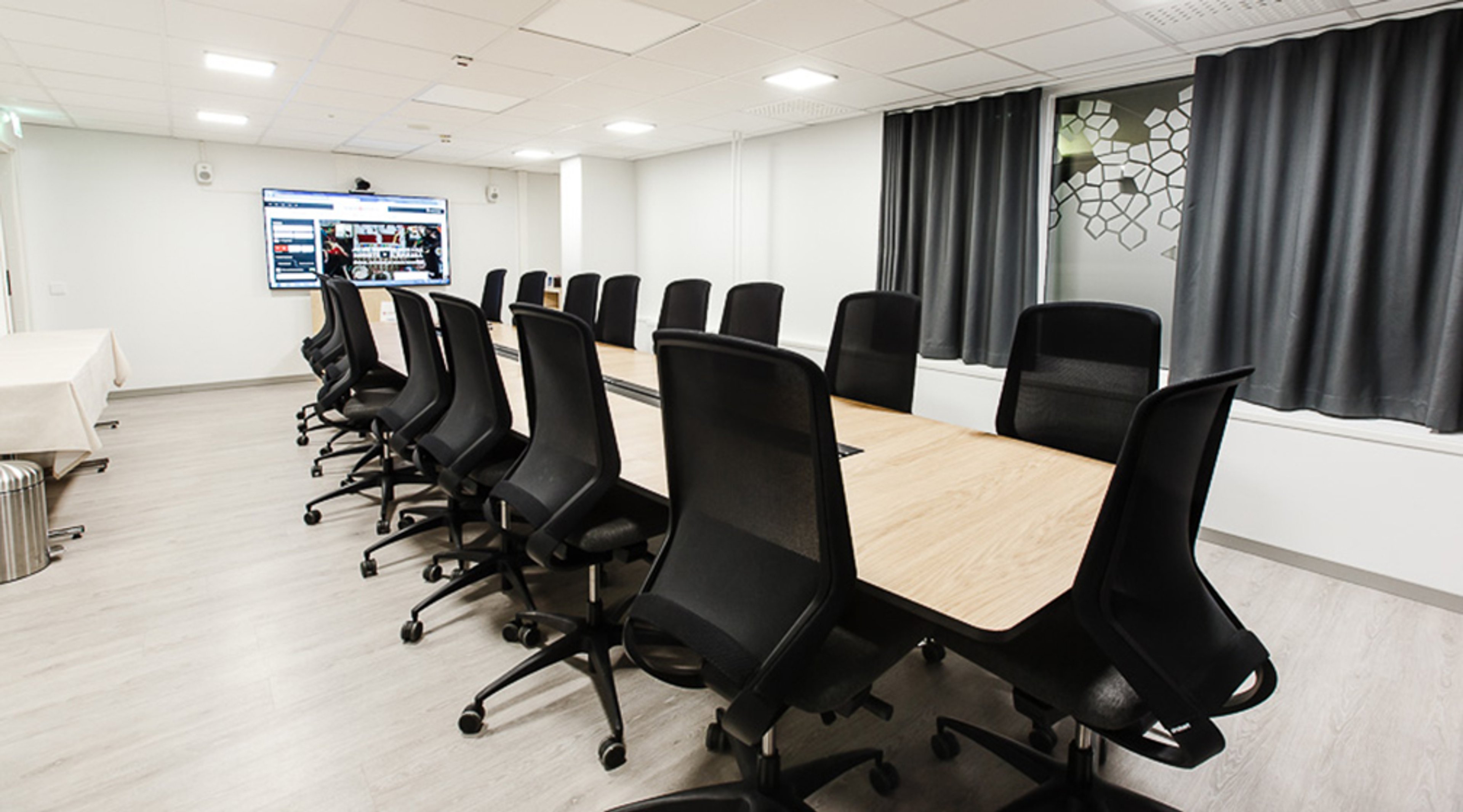 Valoisa neuvotteluhuone, jossa pitkä puinen pöytä ja mustat toimistotuolit. Seinällä iso näyttö ja harmaat verhot. Lattia on vaaleaa laminaattia.