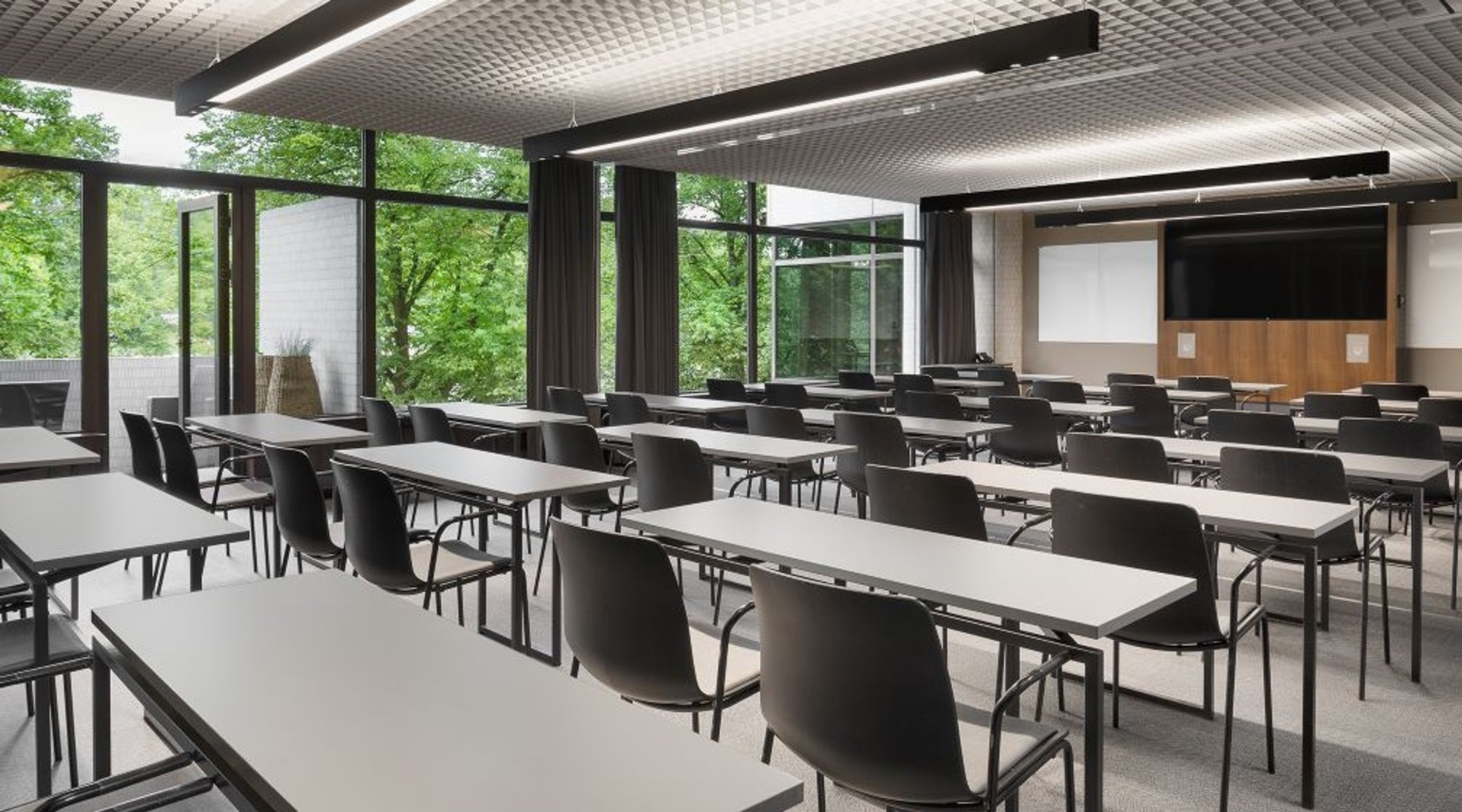 Moderni luokkahuone valkoisilla pöydillä ja mustilla tuoleilla. Suuret ikkunat näyttävät vehreään maisemaan. Edessä projektori ja näyttö.