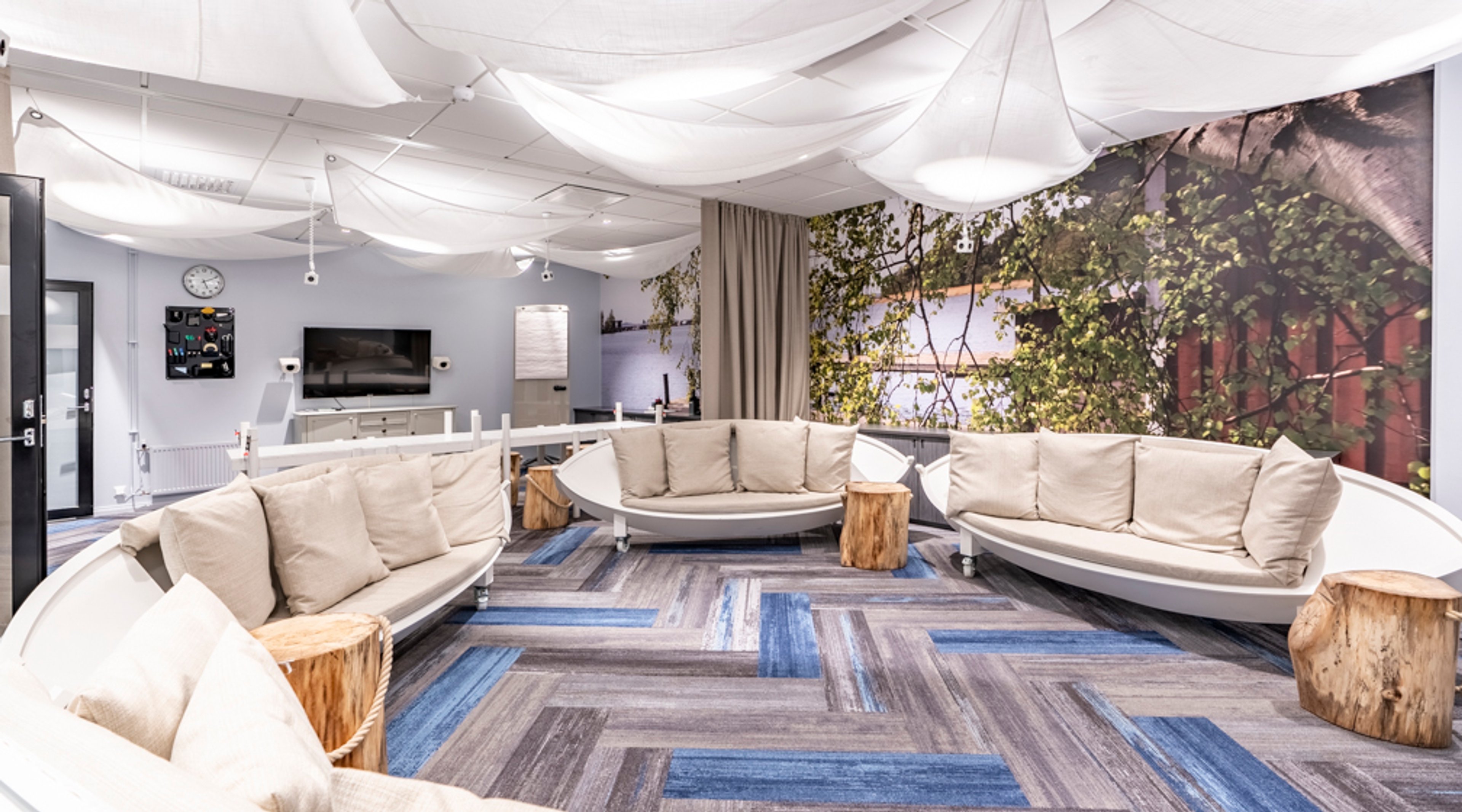 Moderni loungealue, jossa neljä kaartuvaa valkoista sohvaa puisilla jakkaroilla ja sinisävyisellä lattiamatolla. Katossa on valkoisia kangaskatteita.