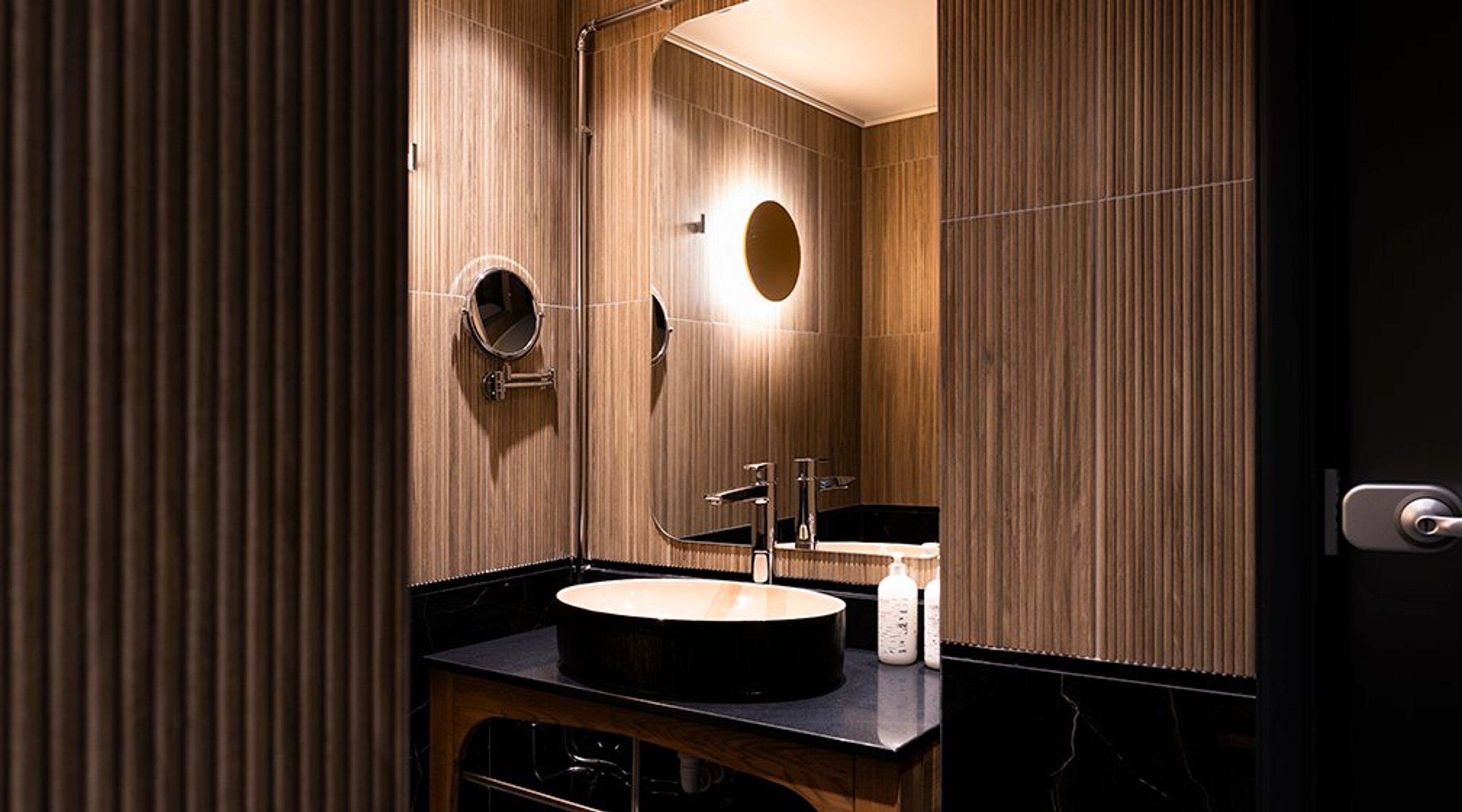 Moderni kylpyhuone puisella paneloinnilla, pyöreällä pesulla, mustalla työtasolla ja valaistulla pyöreällä peilillä.