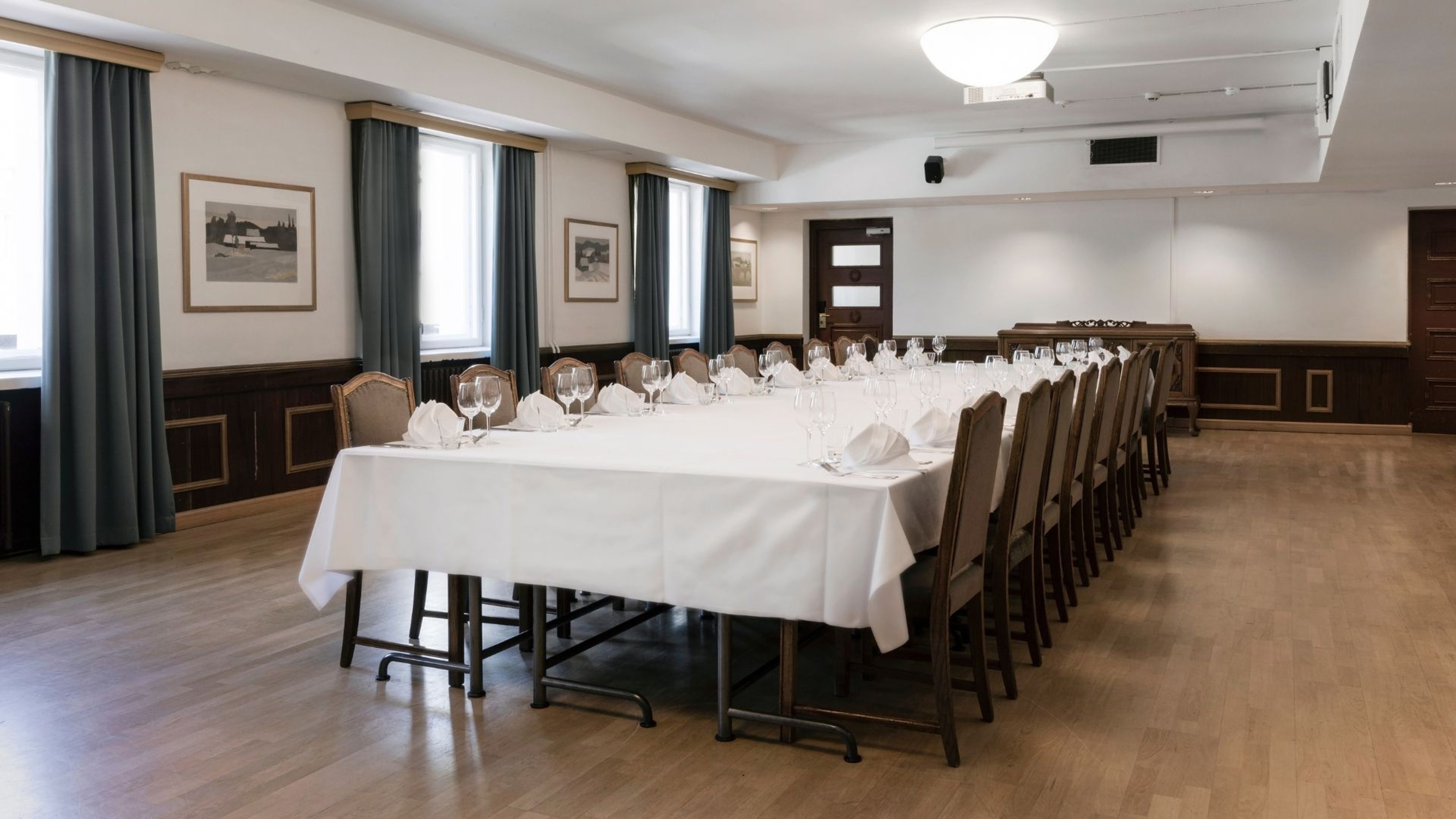 Elegantti kokoushuone, jossa pitkä pöytä katettu valkoisella liinalla ja laseilla. Tilassa puulattia, tummat verhot ja tasainen valaistus.