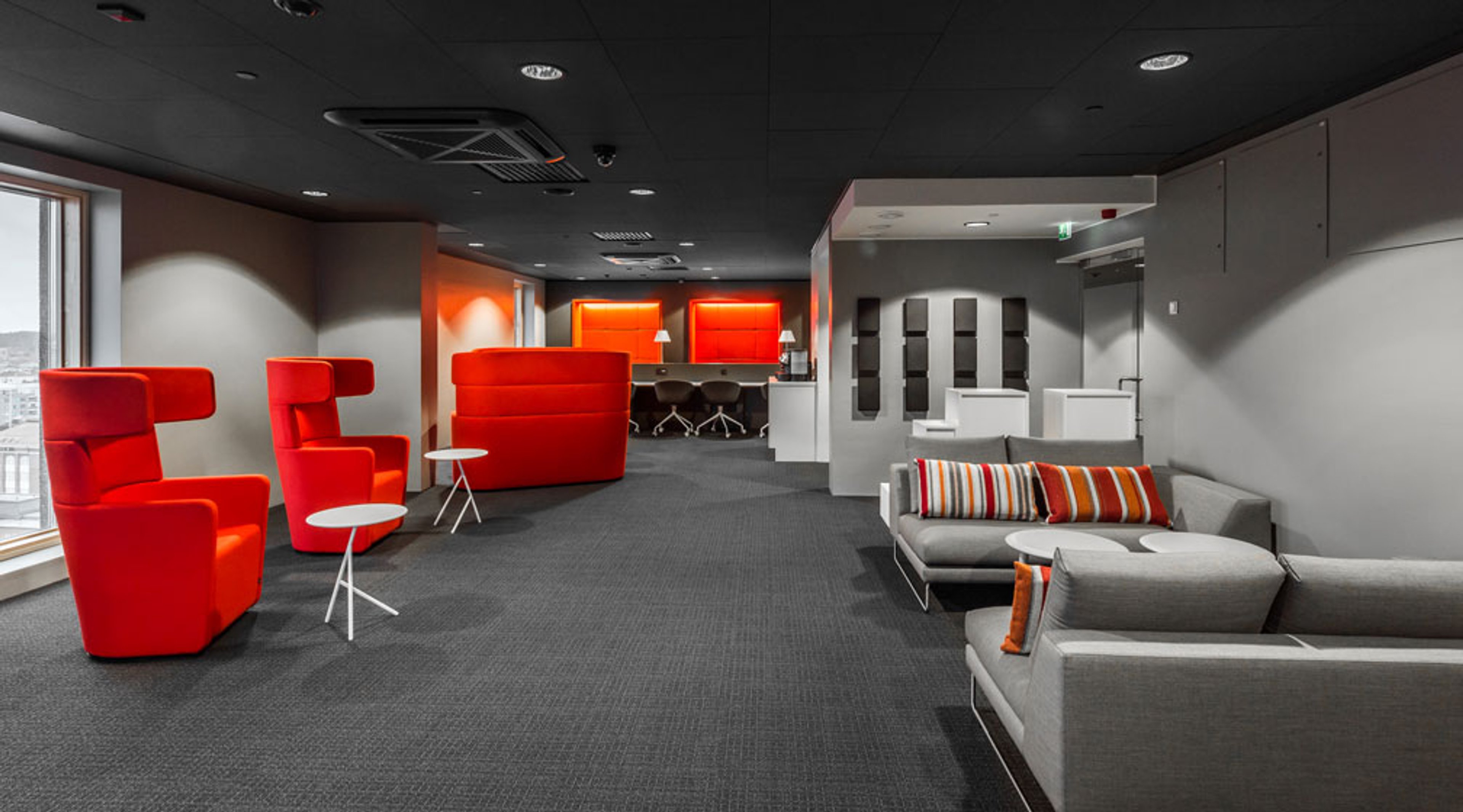 Moderni toimistotila, jossa on punaisia nojatuoleja, harmaita sohvia värikkäillä tyynyillä ja tasainen harmaa kokolattiamatto. Avara lounge-tila isolla ikkunalla.