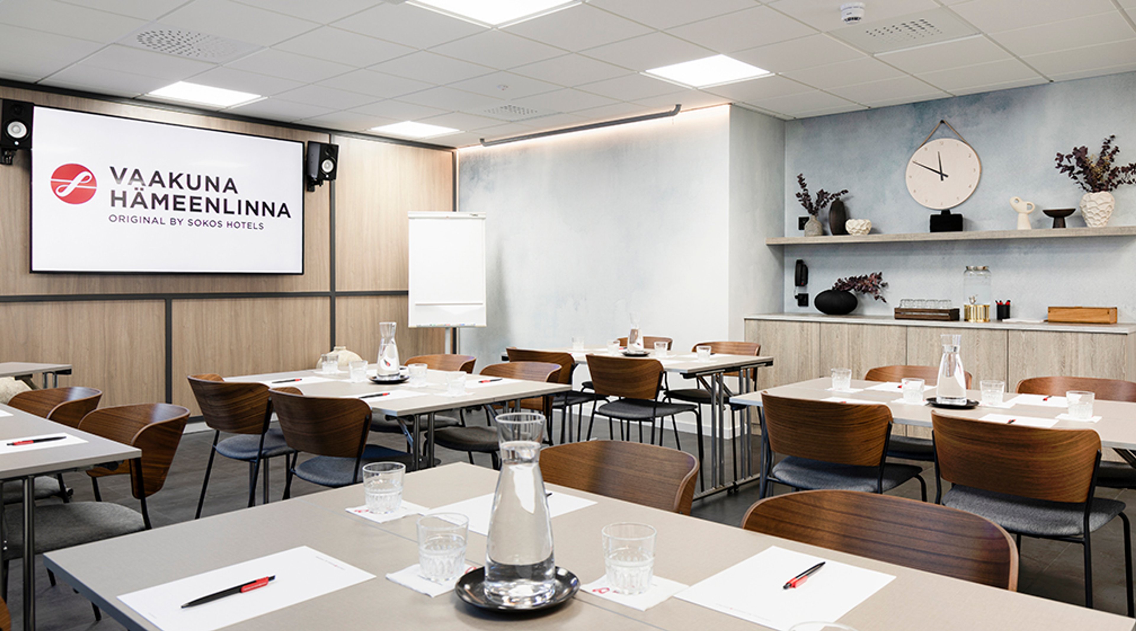 Moderni kokoustila puisilla tuoleilla, pöydillä varustetuilla vesikannuilla ja fläppitaululla. Takaseinällä näytönohjaustauluja ja koriste-esineitä.