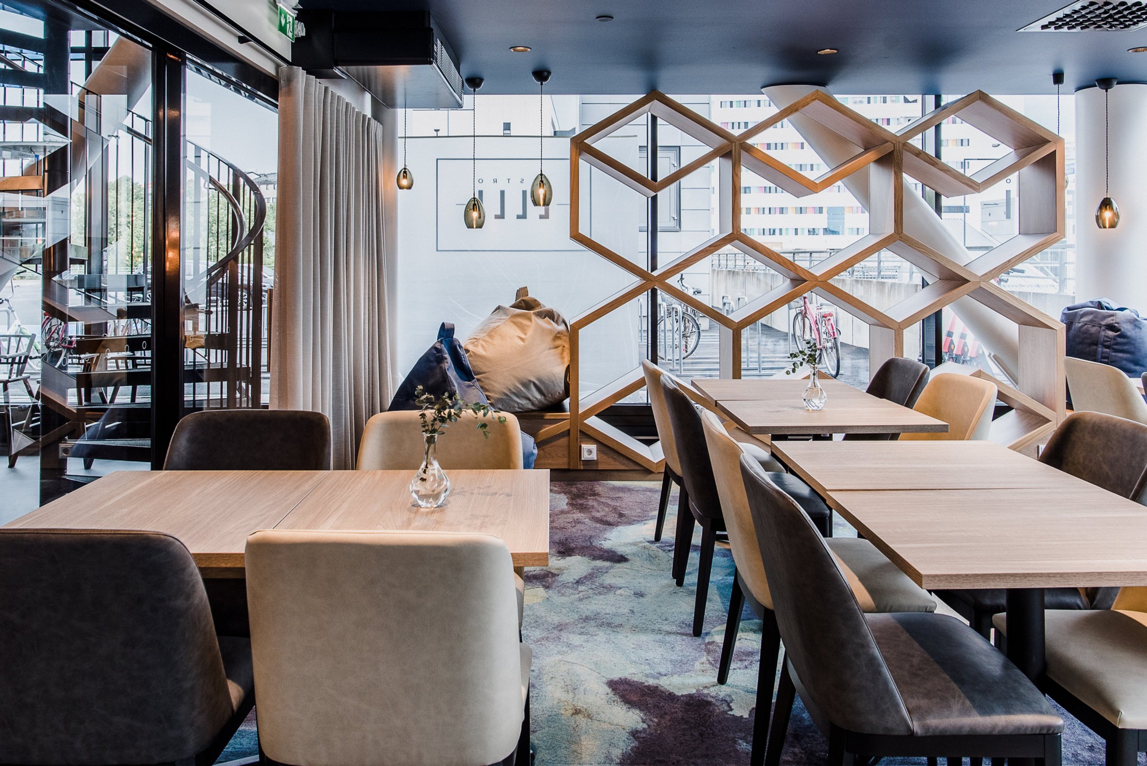 Moderni kahvilatila puisilla pöydillä ja pehmeillä tuoleilla. Kuusikulmioiset puukehykset toimivat tilanjakajina, spiraalinen portaikko vasemmalla.