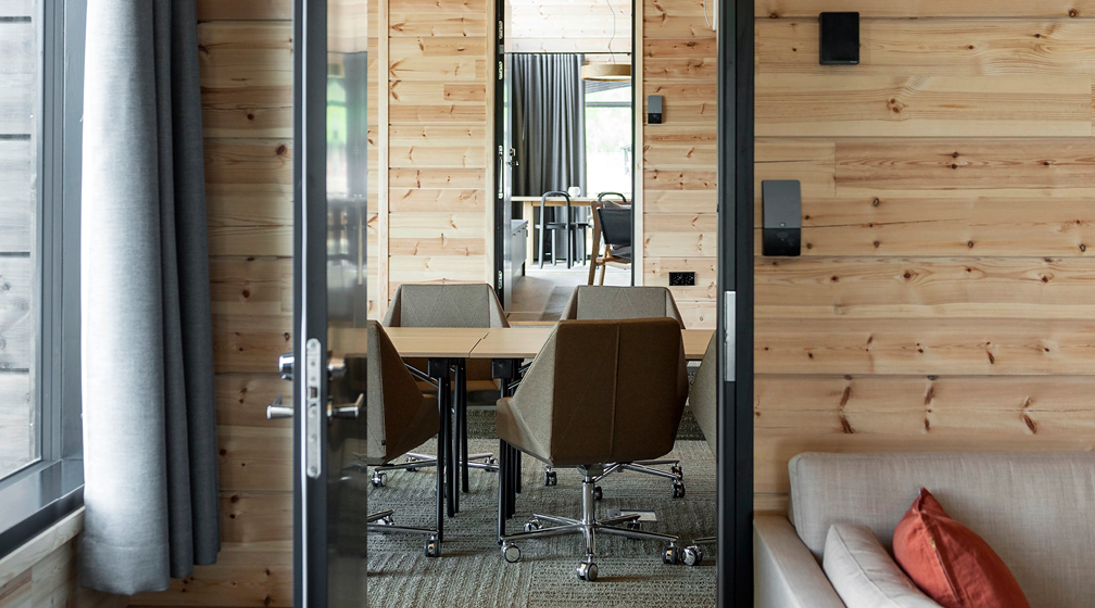 Moderni neuvotteluhuone vaaleaseinäisessä tilassa, jossa puinen pöytä ja neljä harmaata tuolia pyörillä. Laaja oviaukko ja tasainen lattia.