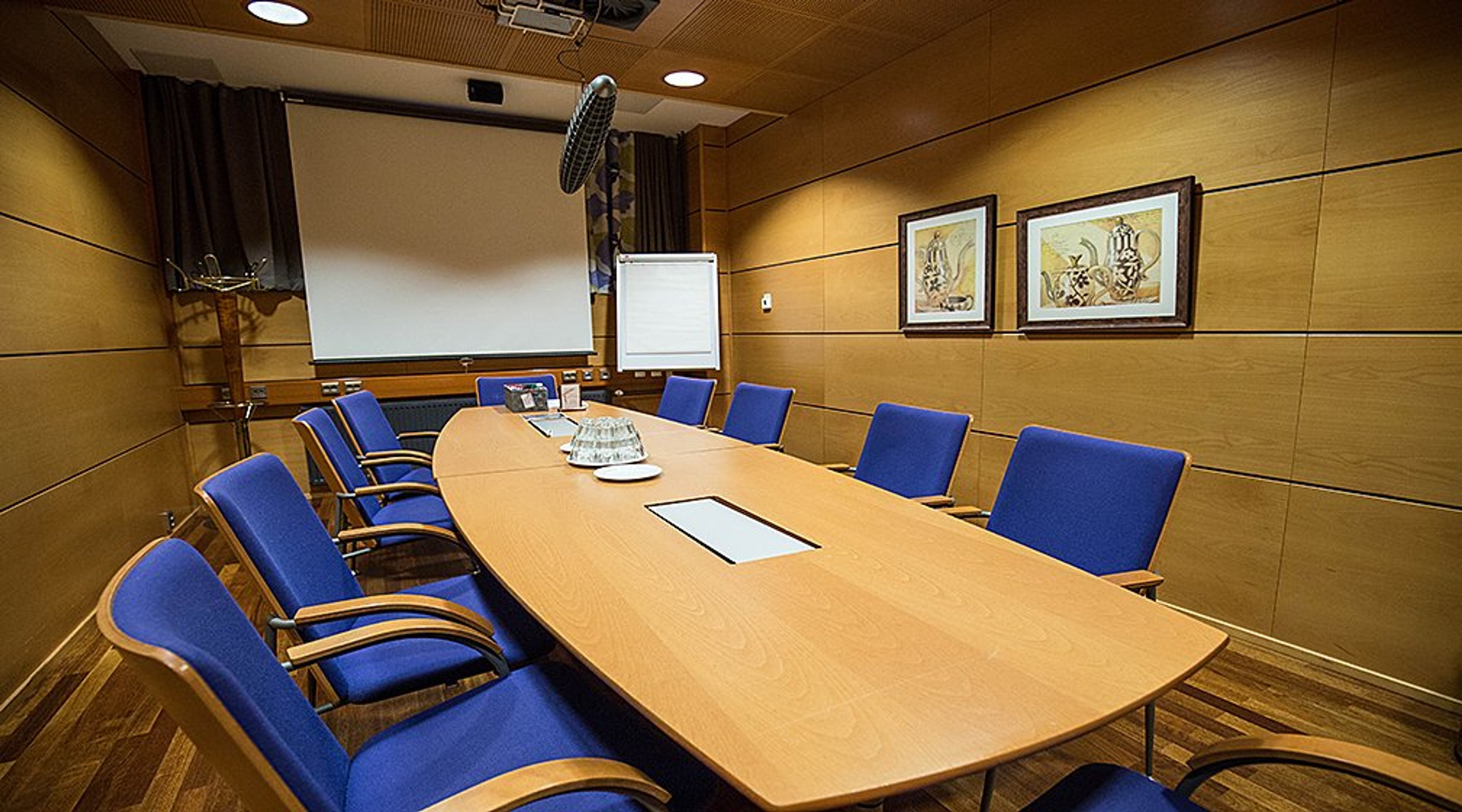 Neuvotteluhuone, jossa on puinen pöytä, siniset tuolit, valkokangas ja fläppitaulu. Puupaneloidut seinät ja kaksi taulua.