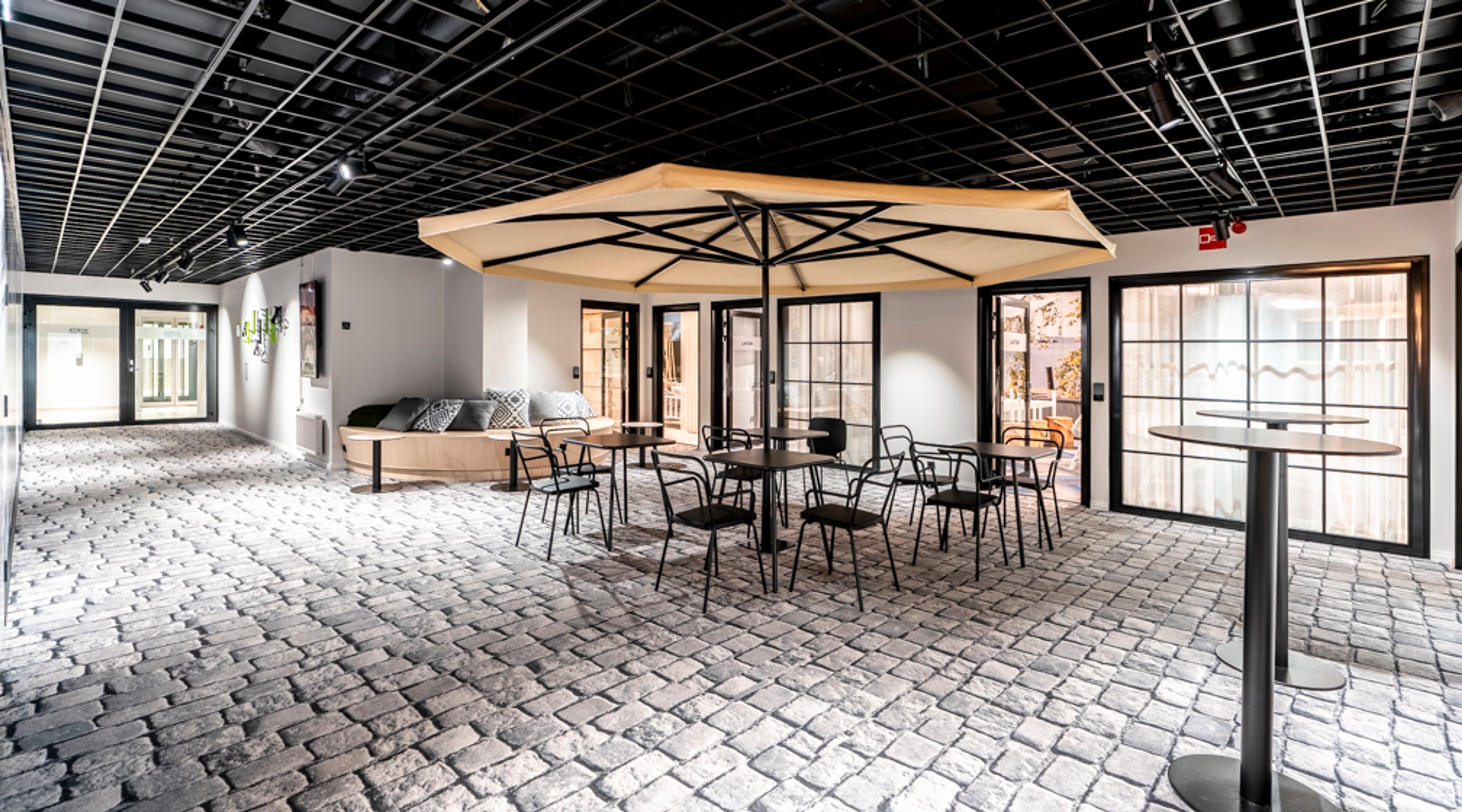 Sisätila, jossa kivilaattalattia, suuri terassivarjo pöytäryhmän yllä ja lasiseinät. Avara tila mustalla kattoritilällä ja lepoalueella.