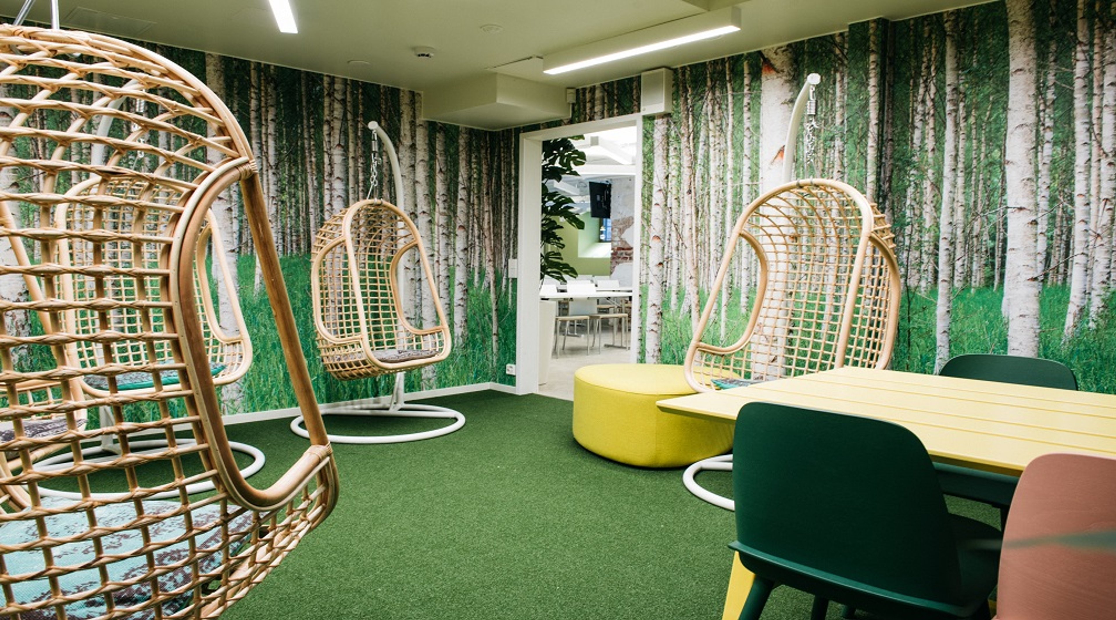 Rentoutumistila, jossa riippuvia rottinkikeinuja, metsäaiheisia seinäkuvia, vihreä kokolattiamatto ja pöytäryhmä. Vaalea valaistus ja avoin pääsy viereiseen tilaan.