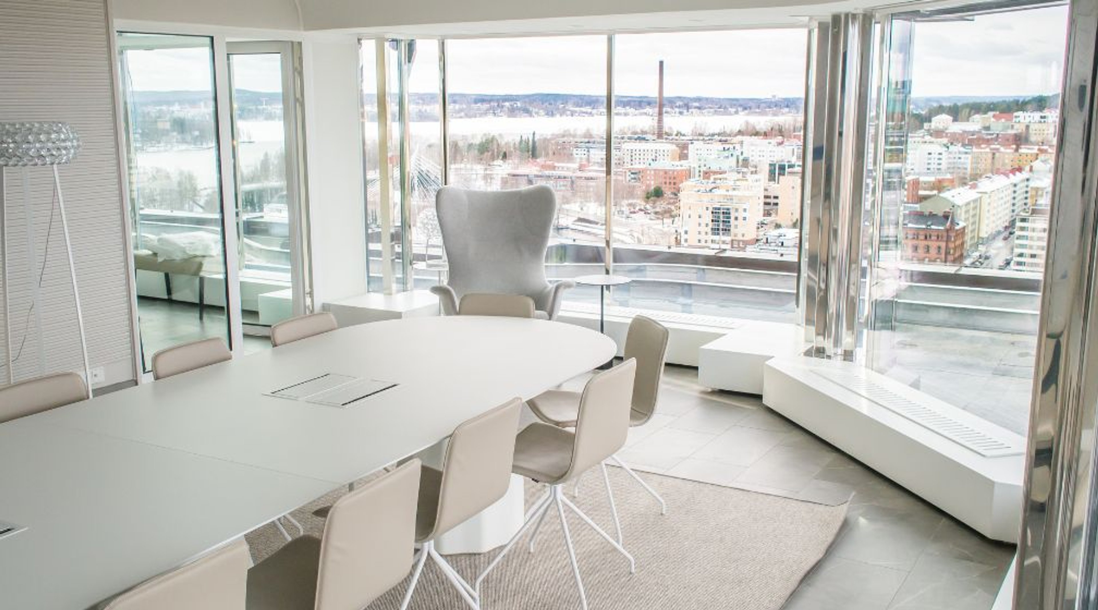 Moderni neuvotteluhuone valkoisella pöydällä, tuoleilla ja nojatuolilla. Suurista ikkunoista avautuu näkymä kaupunkiin.