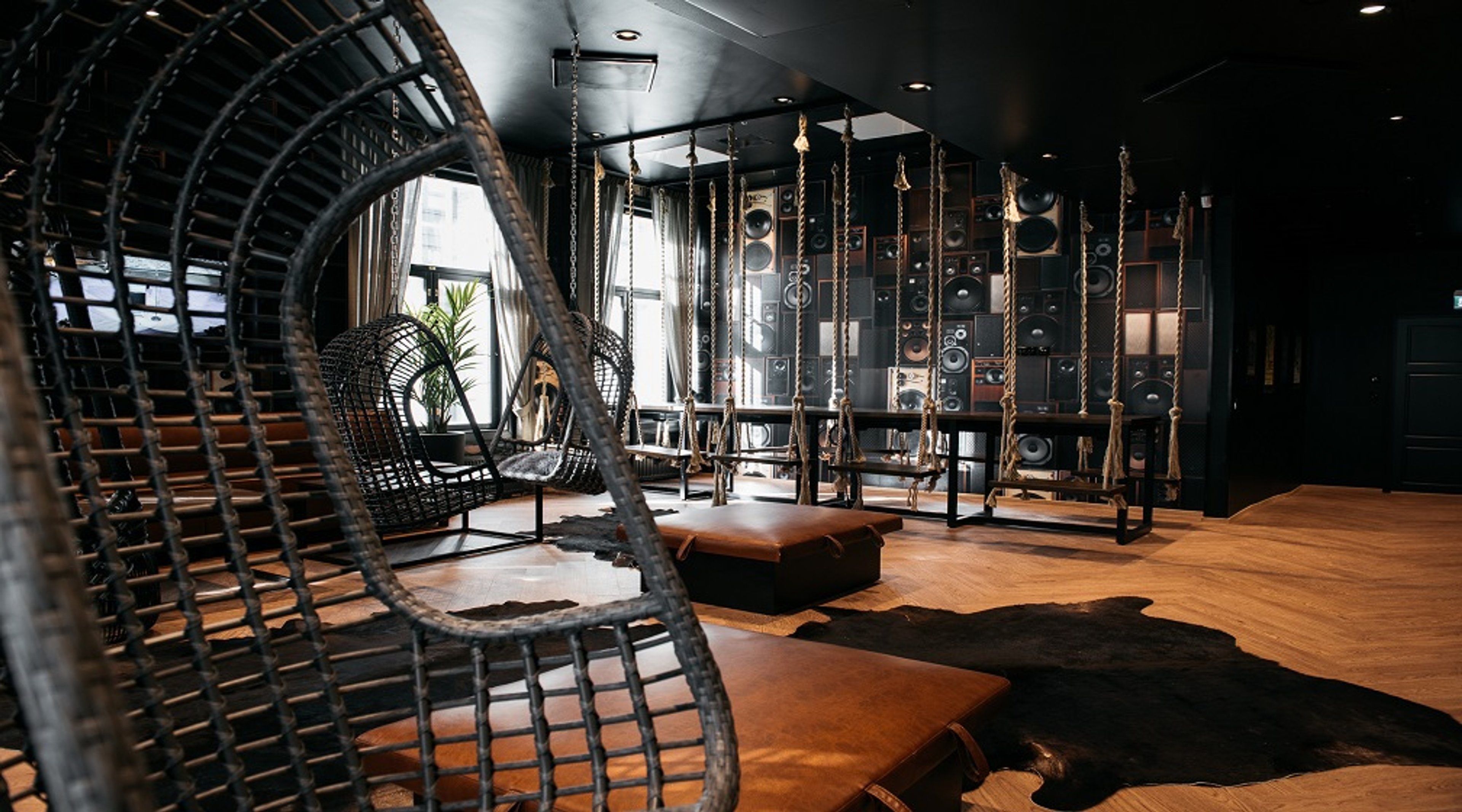 Tumma, moderni huone jossa riippuvia korijakkaroita, nahkaistuimia ja kaapista tehtyjä kaiutinseiniä. Puulattia ja eläintalja mattoina.