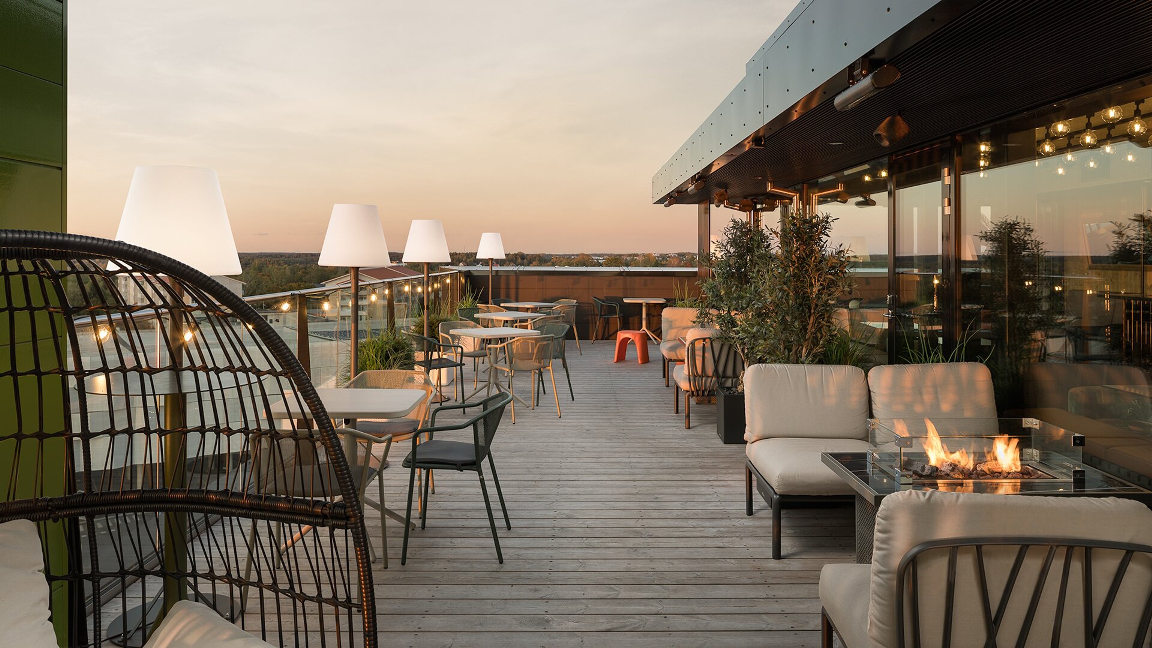 Kattoterassi auringonlaskun aikaan, puulattialla on pöytiä, tuoleja ja sohvia. Oikealla takka, ympärillä kasveja ja suuret valaisimet.