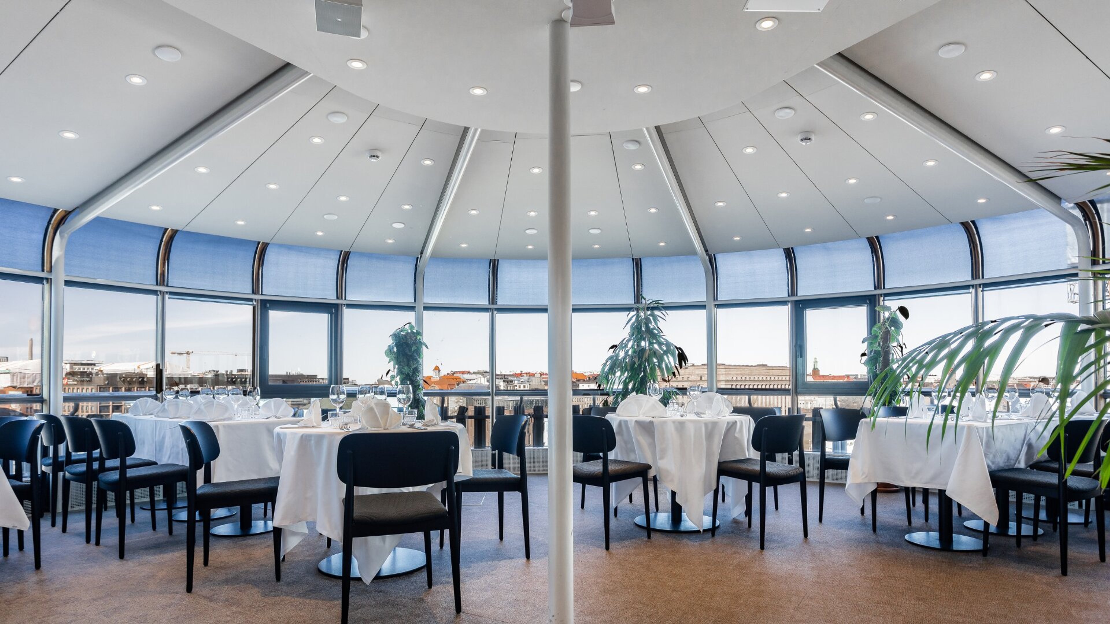 Pyöreä ravintolasali panoraamaikkunoilla. Valkoisten pöytäliinojen peittämät pöydät ja mustat tuolit, taustalla kaupunkimaisema. Tasainen lattia, runsas valaistus.