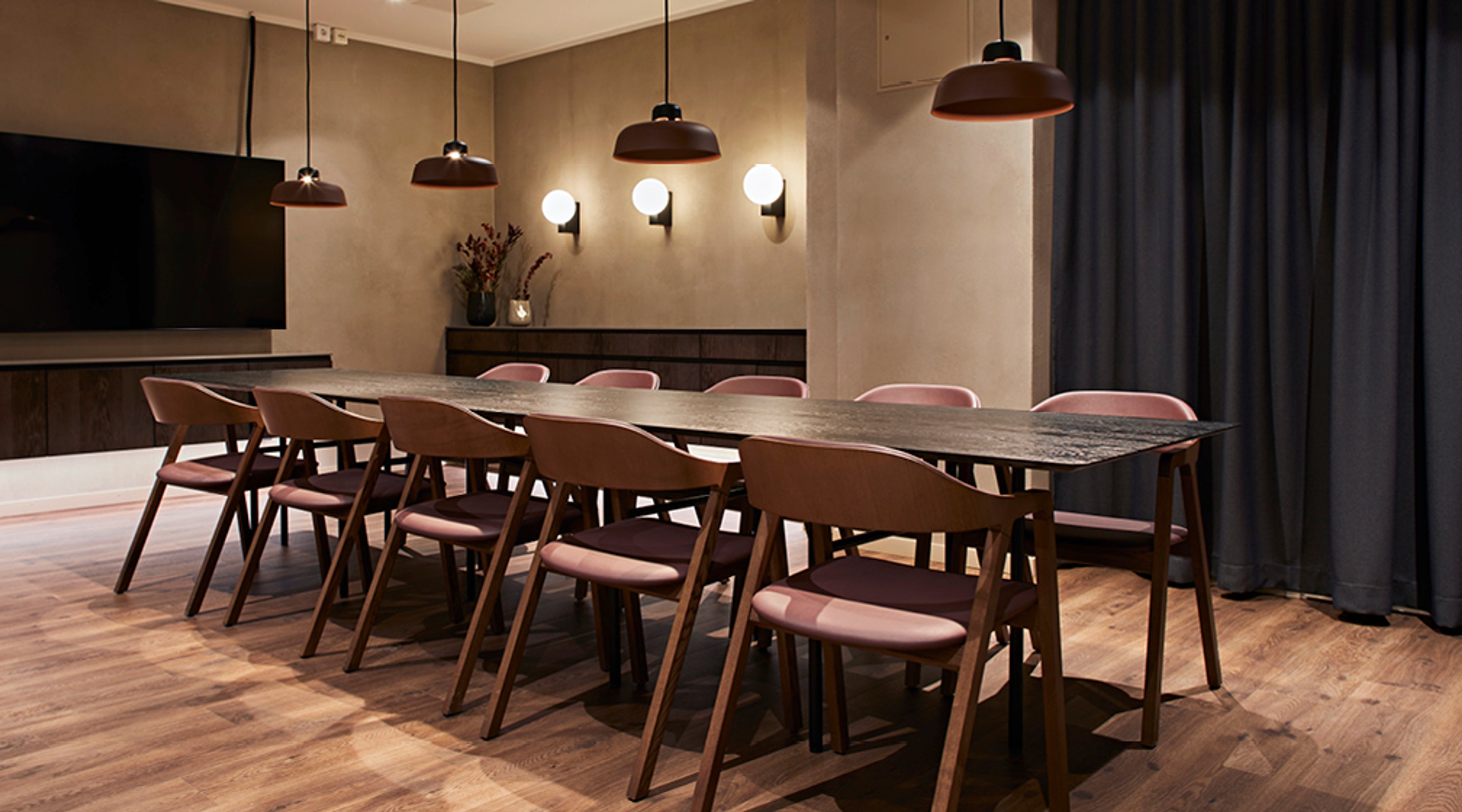 Moderni kokoushuone pitkällä puupöydällä, vaaleanpunaisilla tuoleilla, riippuvalaisimilla ja seinävalaisimilla. Puulattia ja tumma verho.