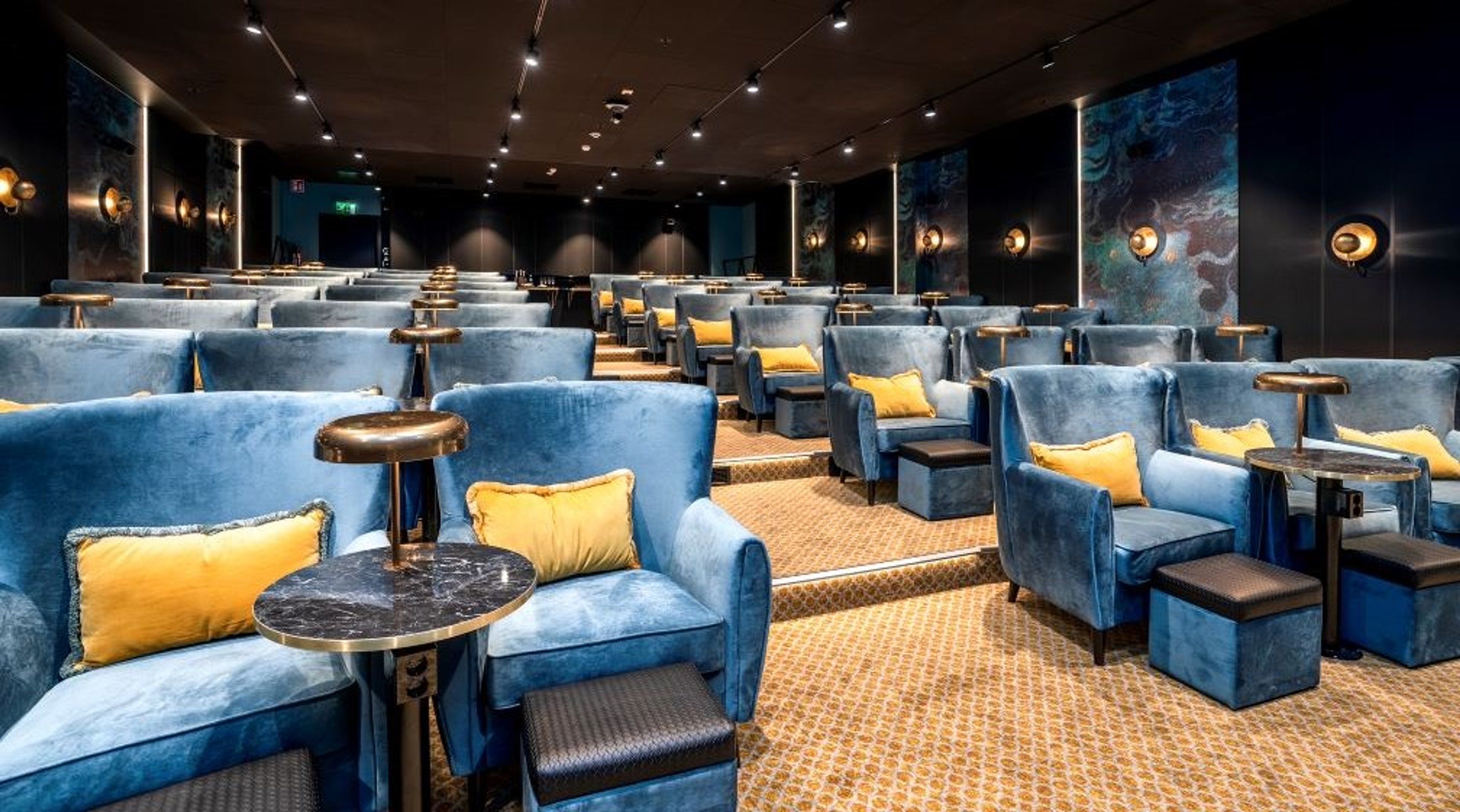 Ylellinen auditoriotila sinisin nojatuolein ja keltaisin tyynyin. Pienikokoiset marmoripöydät istuinten välissä. Seinät tummat ja valaistu kohdevaloilla.