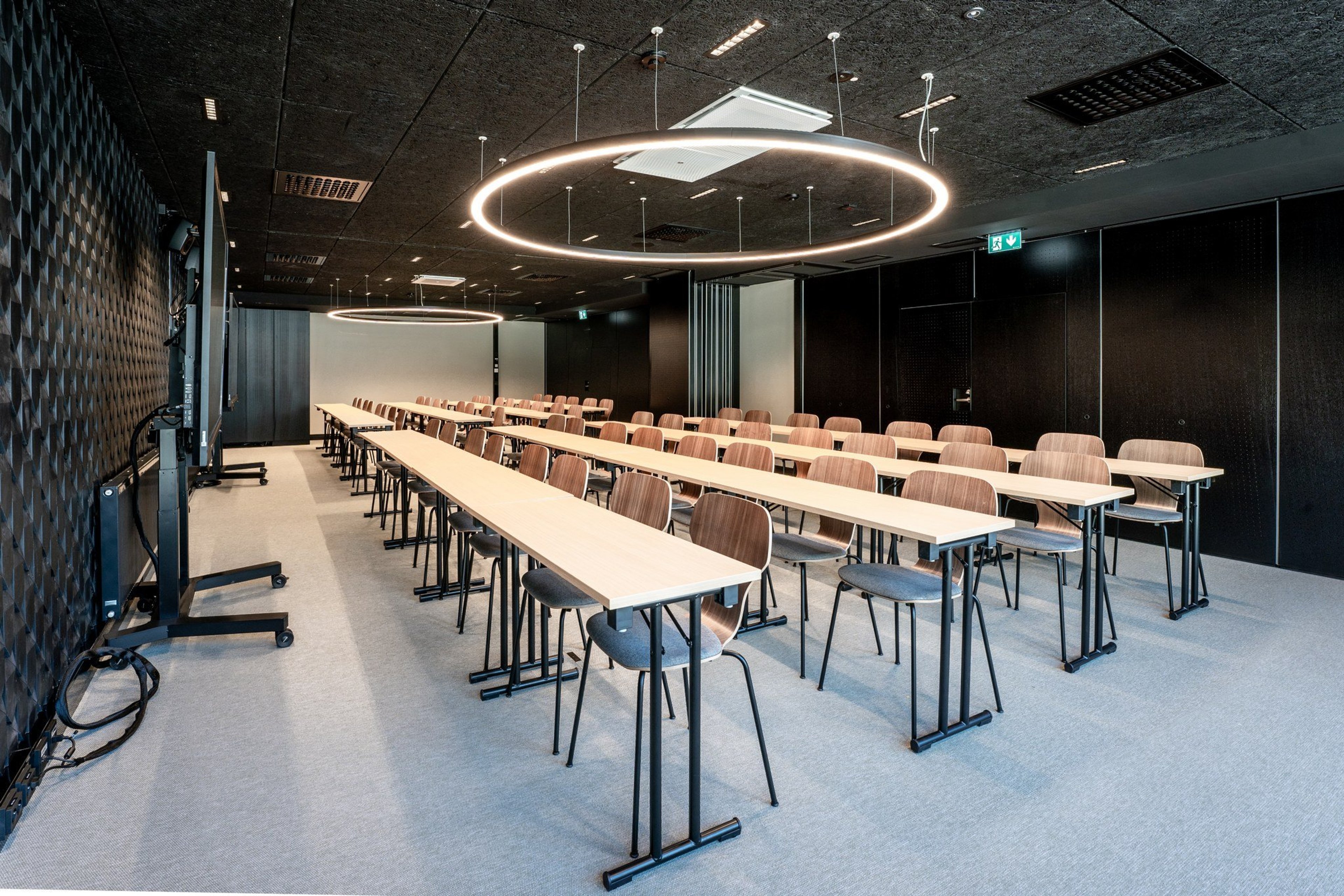 Moderni kokoustila, jossa pitkiä pöytiä ja puutuoleja, katossa rengasmaiset valaisimet, tasainen lattiapinta ja tummat seinät.
