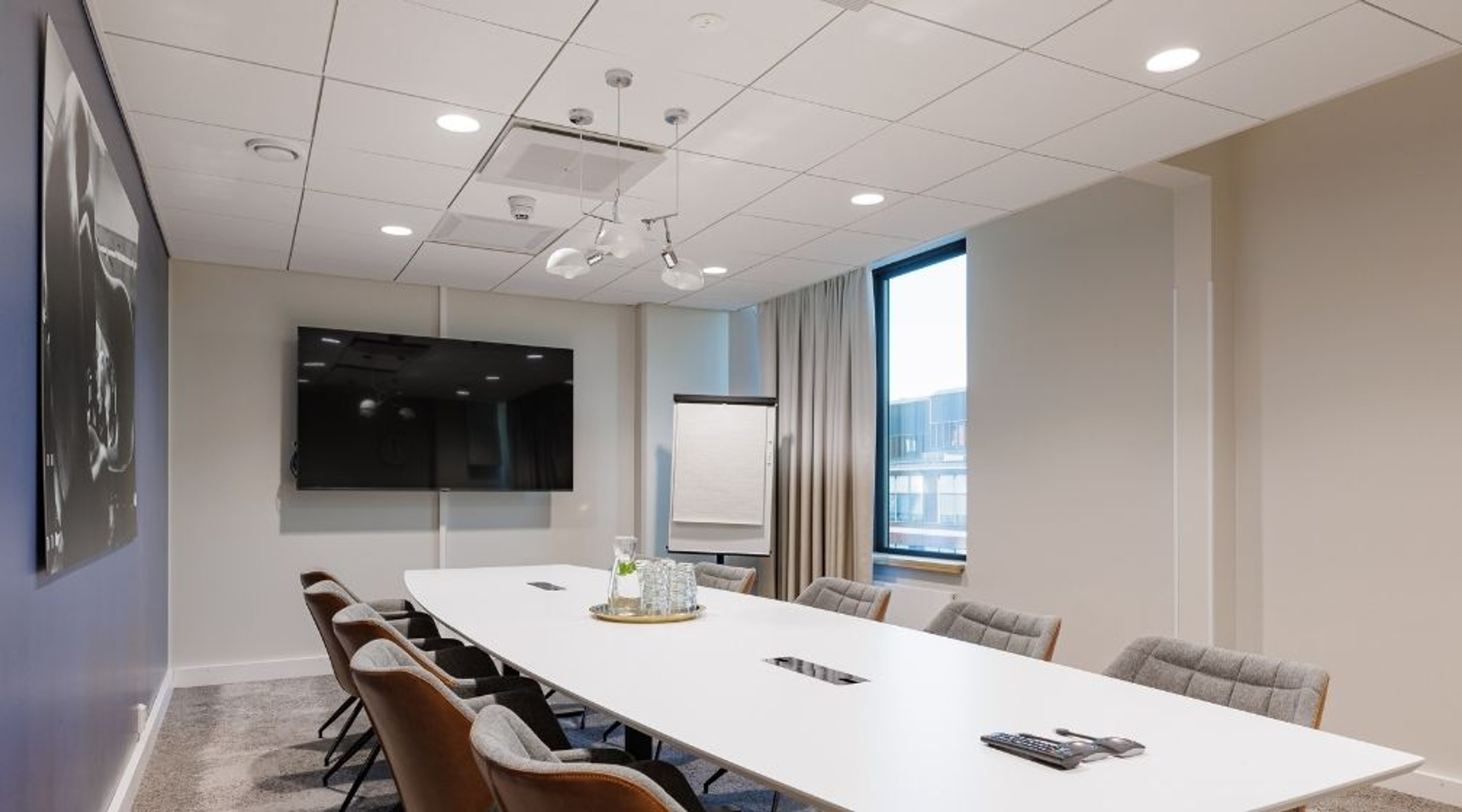 Moderni neuvotteluhuone, jossa pitkä valkoinen pöytä, mukavat tuolit, suuri näyttö seinällä ja fläppitaulu. Valoisa tila isolla ikkunalla.