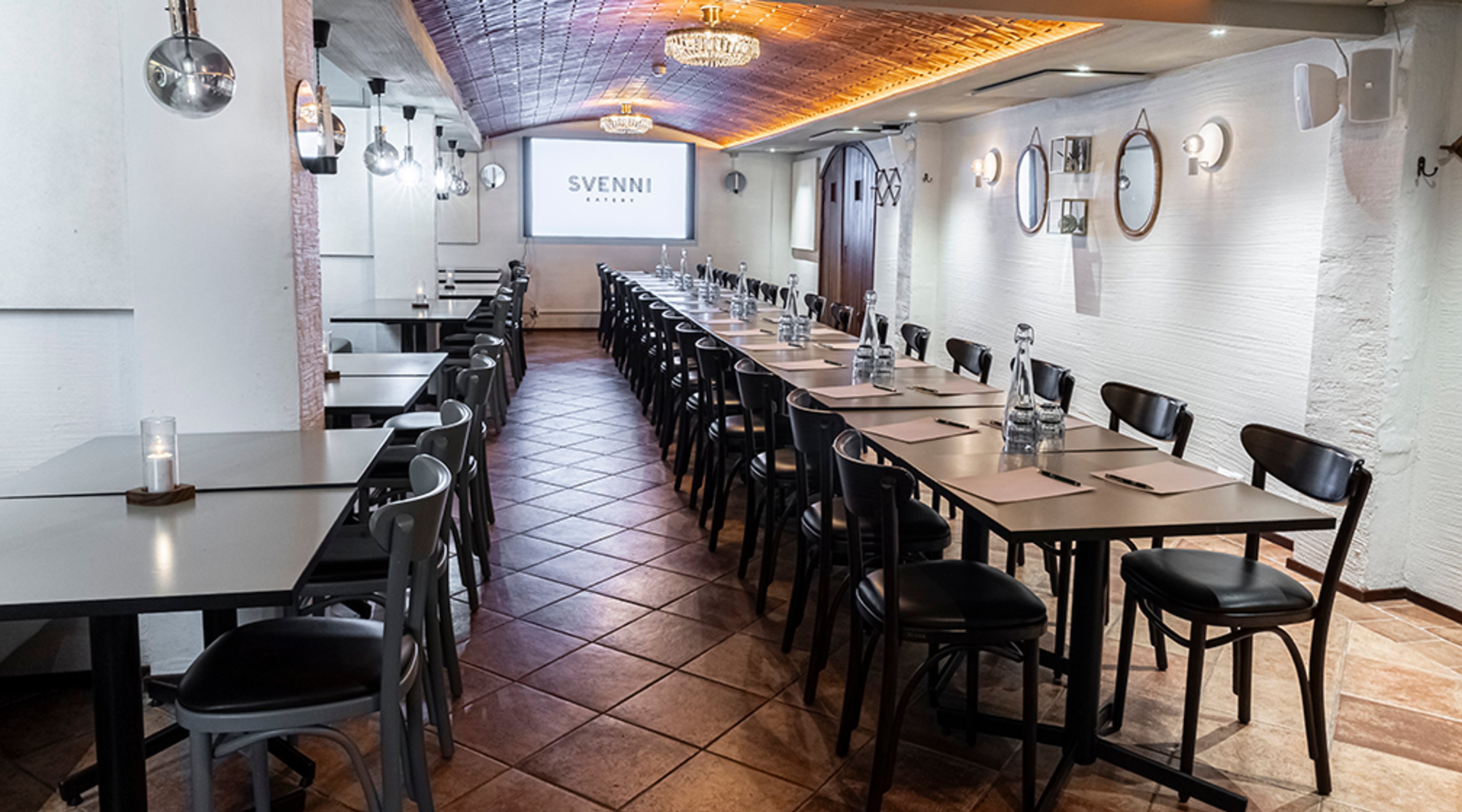 Pitkä kokoushuone, jossa ruskea laattalattia ja pitkä pöytäjärjestys. Valkoinen tiiliseinä, valkokangas ja koristeelliset kattovalaisimet.