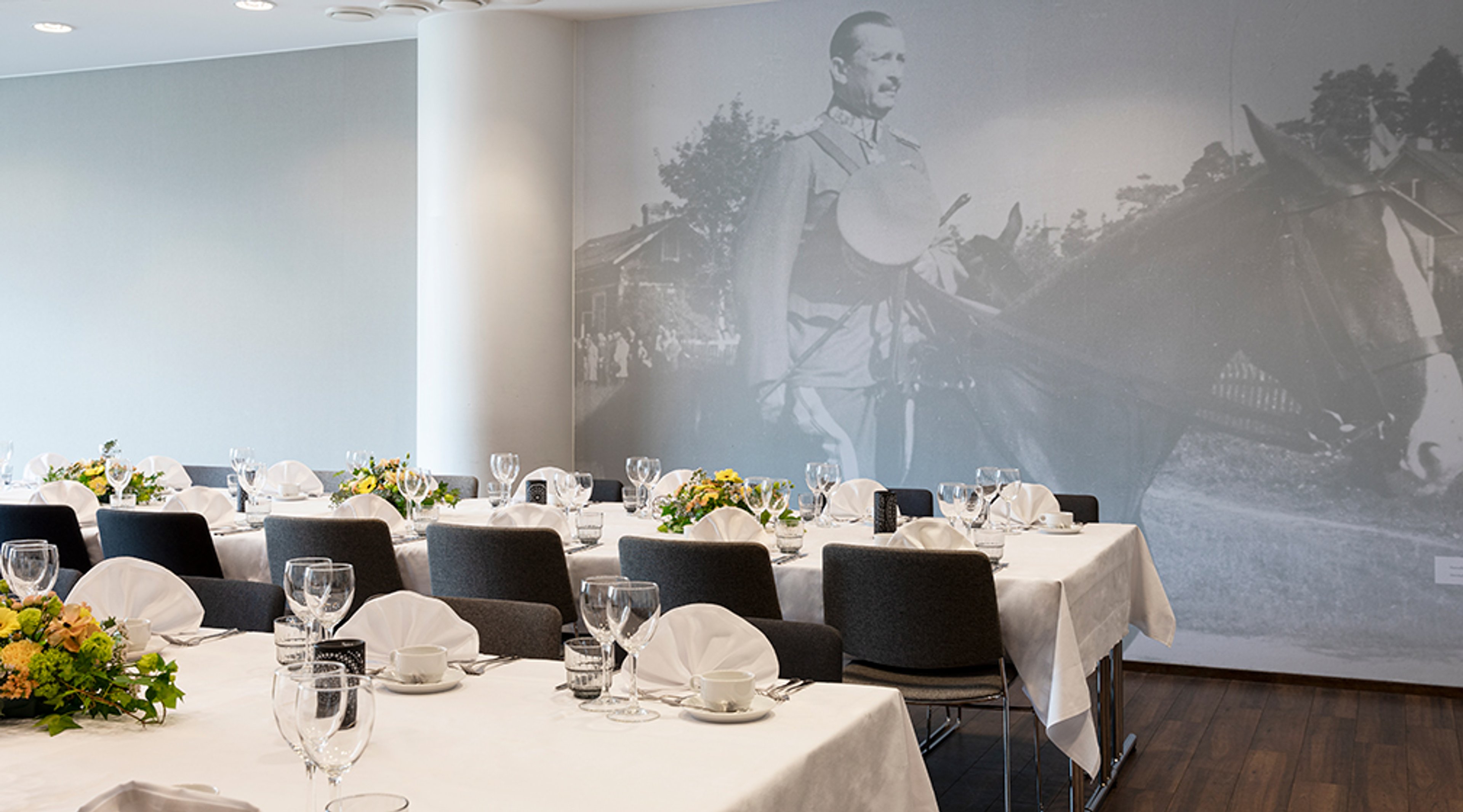 Juhlatila, jossa valkoisilla pöytäliinoilla katetut pöydät, mustat tuolit ja seinällä historiallinen mustavalkoinen valokuva. Puulattia.