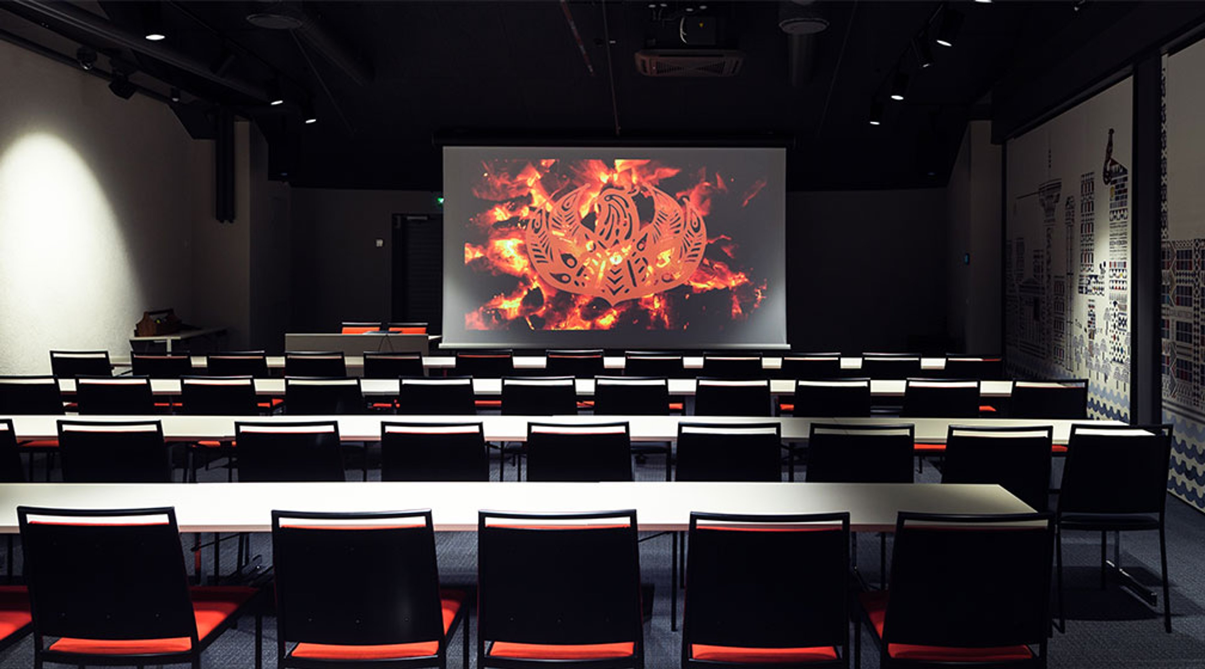 Tumma auditoriotila, jossa rivejä pöytiä ja tuoleja punaisten istuintyynyjen kera. Valkokankaalla näkyy tulinen taidekuva.