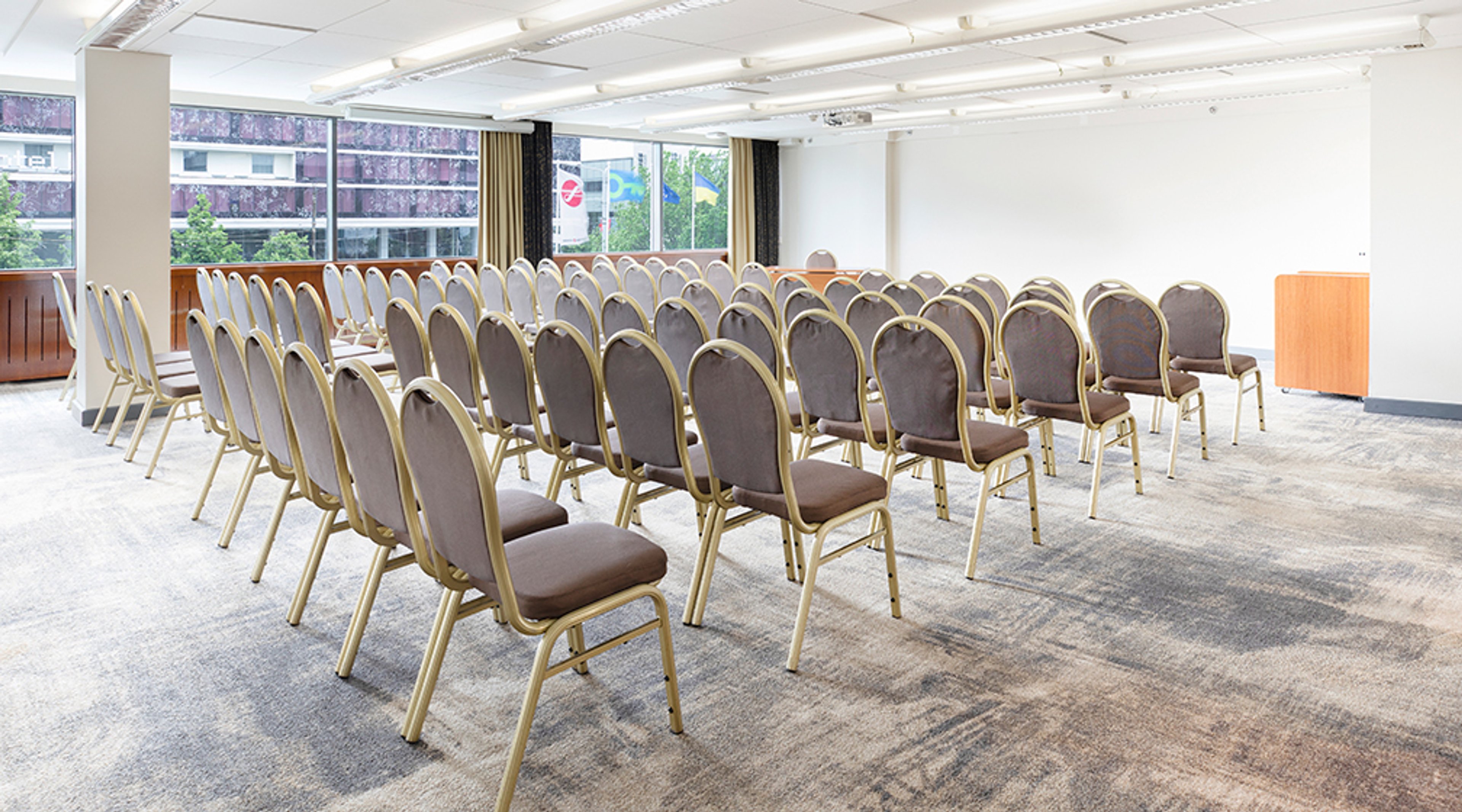 Bright conference room with rows of grey conference chairs with gold-coloured frames, low-pile carpet and large windows.