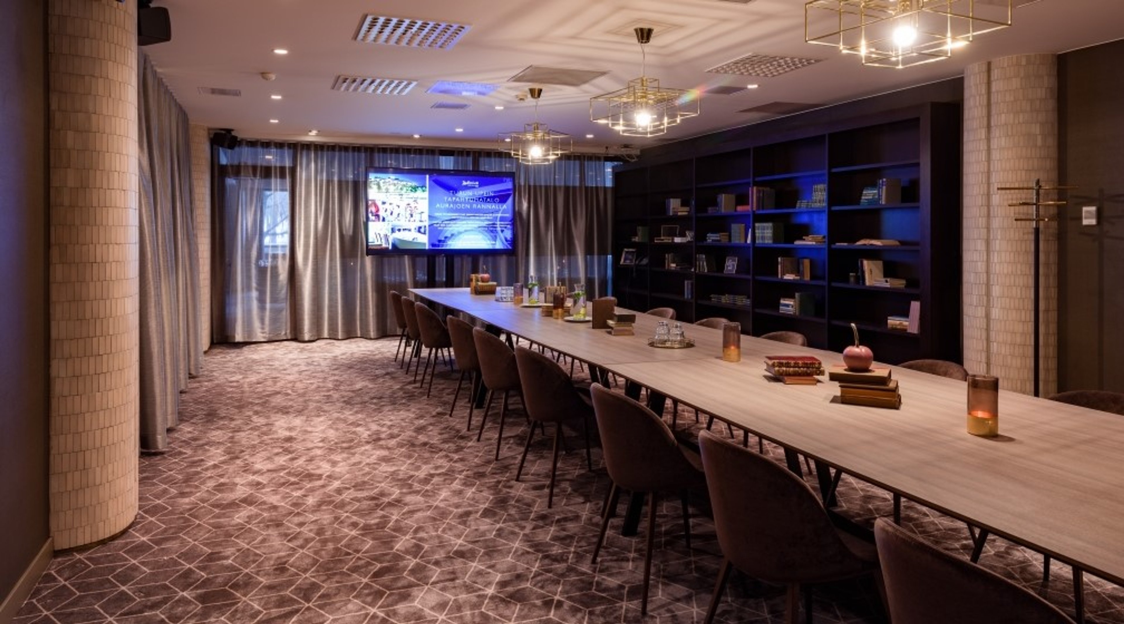 Kokoushuone pitkällä puupöydällä, tuoleilla, kirjahyllyillä ja suurella näytöllä seinällä. Hillitty valaistus ja kuviollinen lattia.