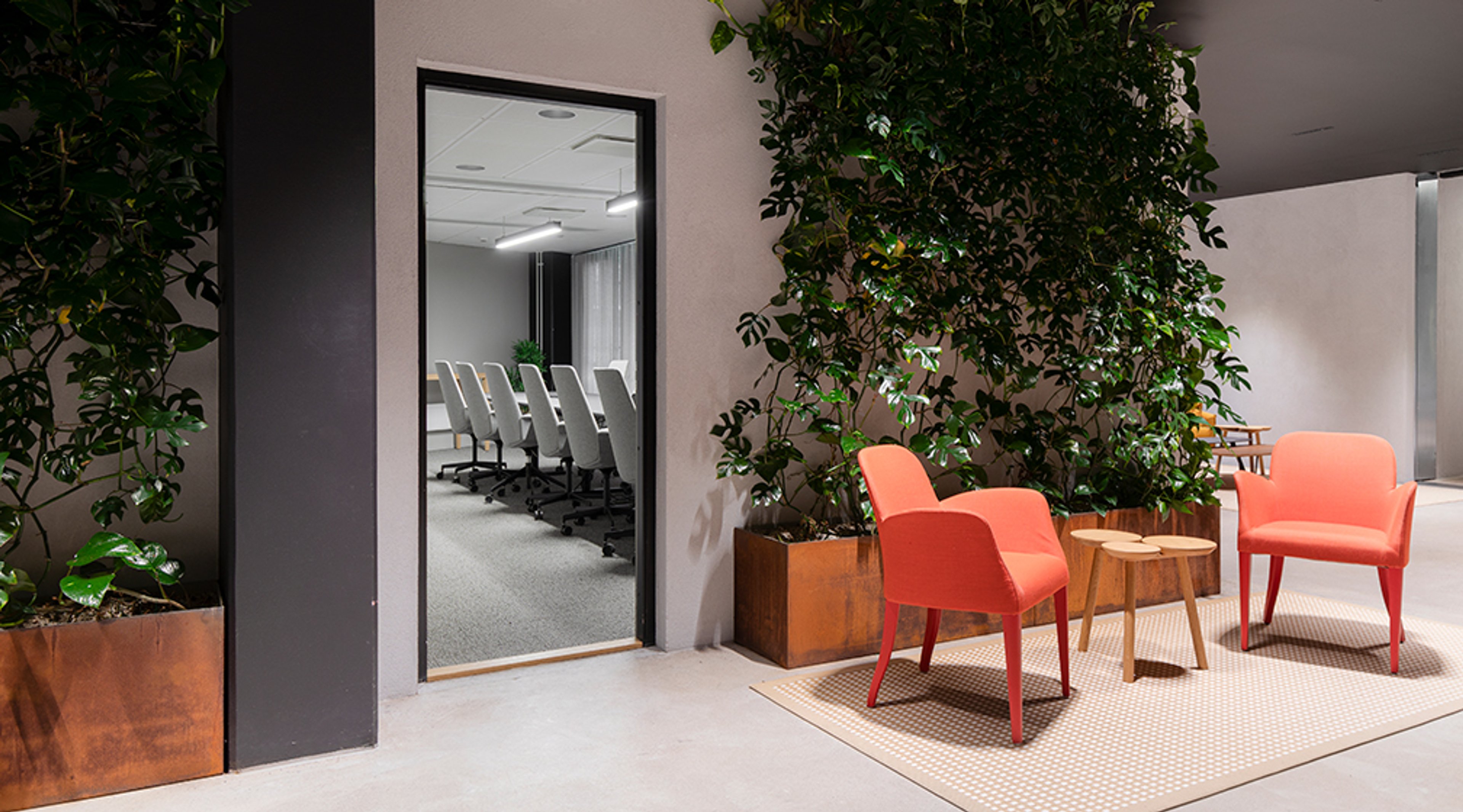 Modern office waiting area with orange chairs and indoor plants. An open doorway leads to a meeting room with white chairs.
