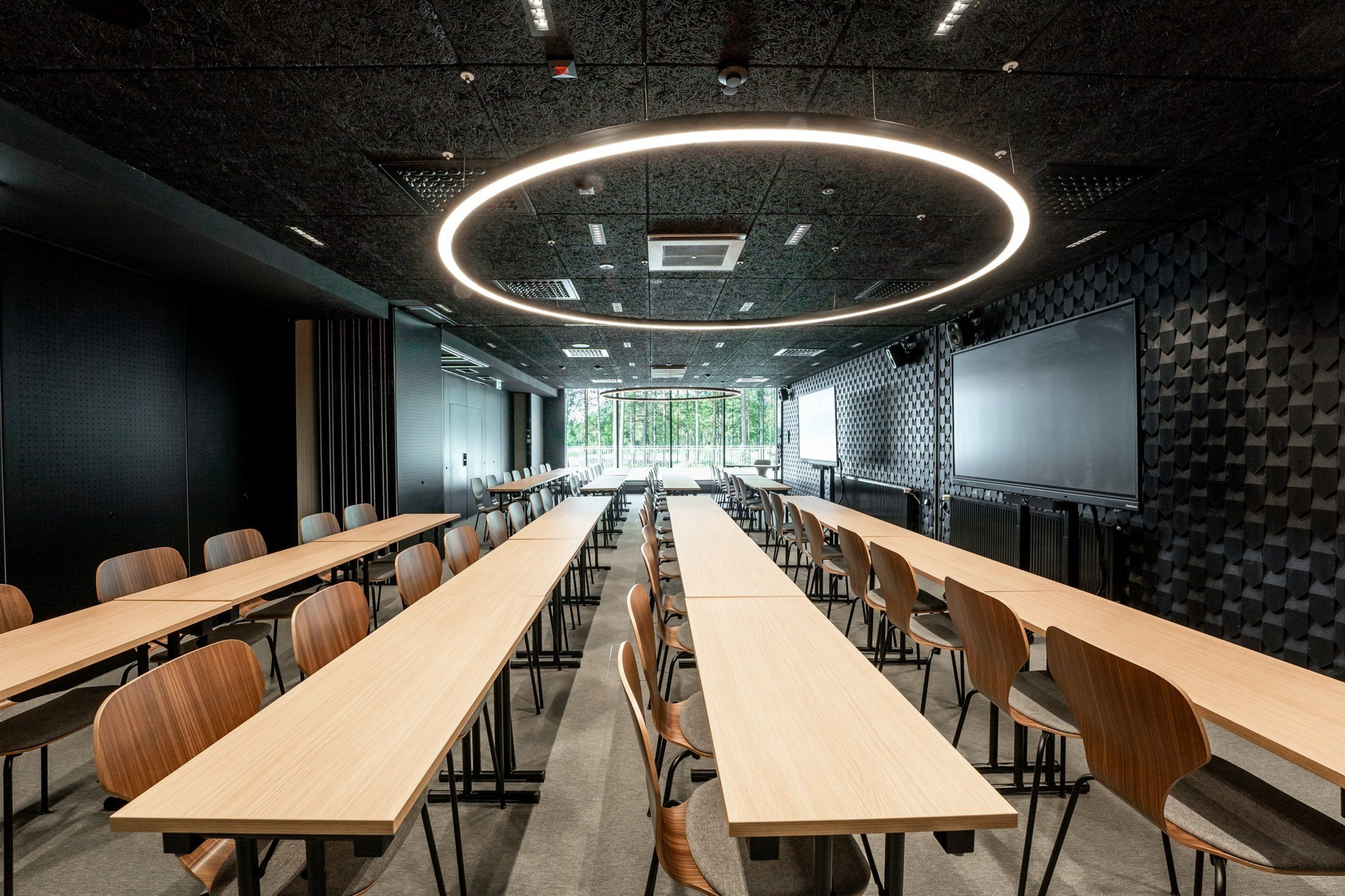 Moderni kokoustila, jossa puisia pöytiä riveissä, katossa pyöreä valorengas, seinillä näytöt ja takana isot ikkunat luontonäkymällä.