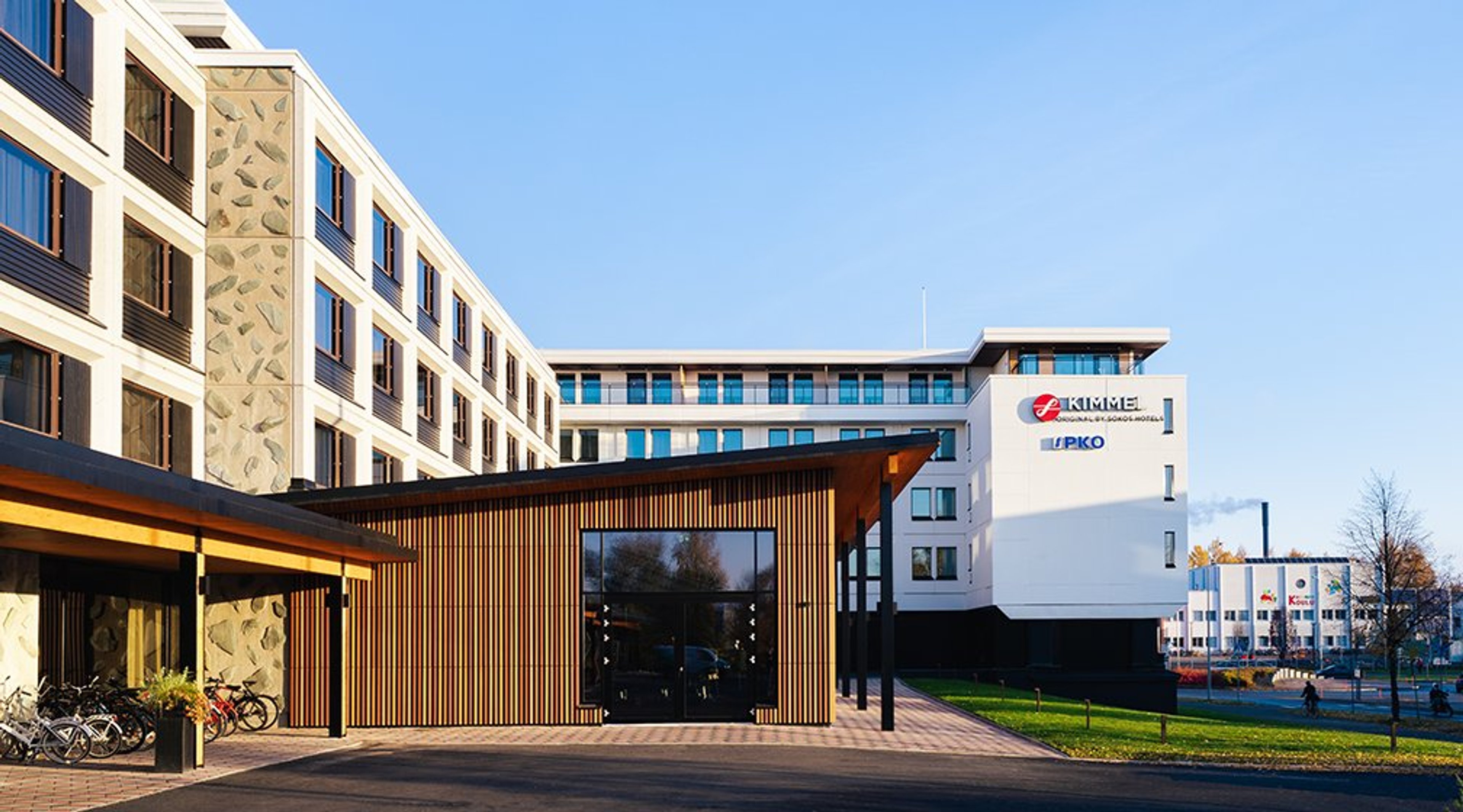 Moderni valkoinen toimistorakennus puisella sisäänkäynnillä. Rakennuksen edessä pyörätelineet ja tasainen kävelytie.