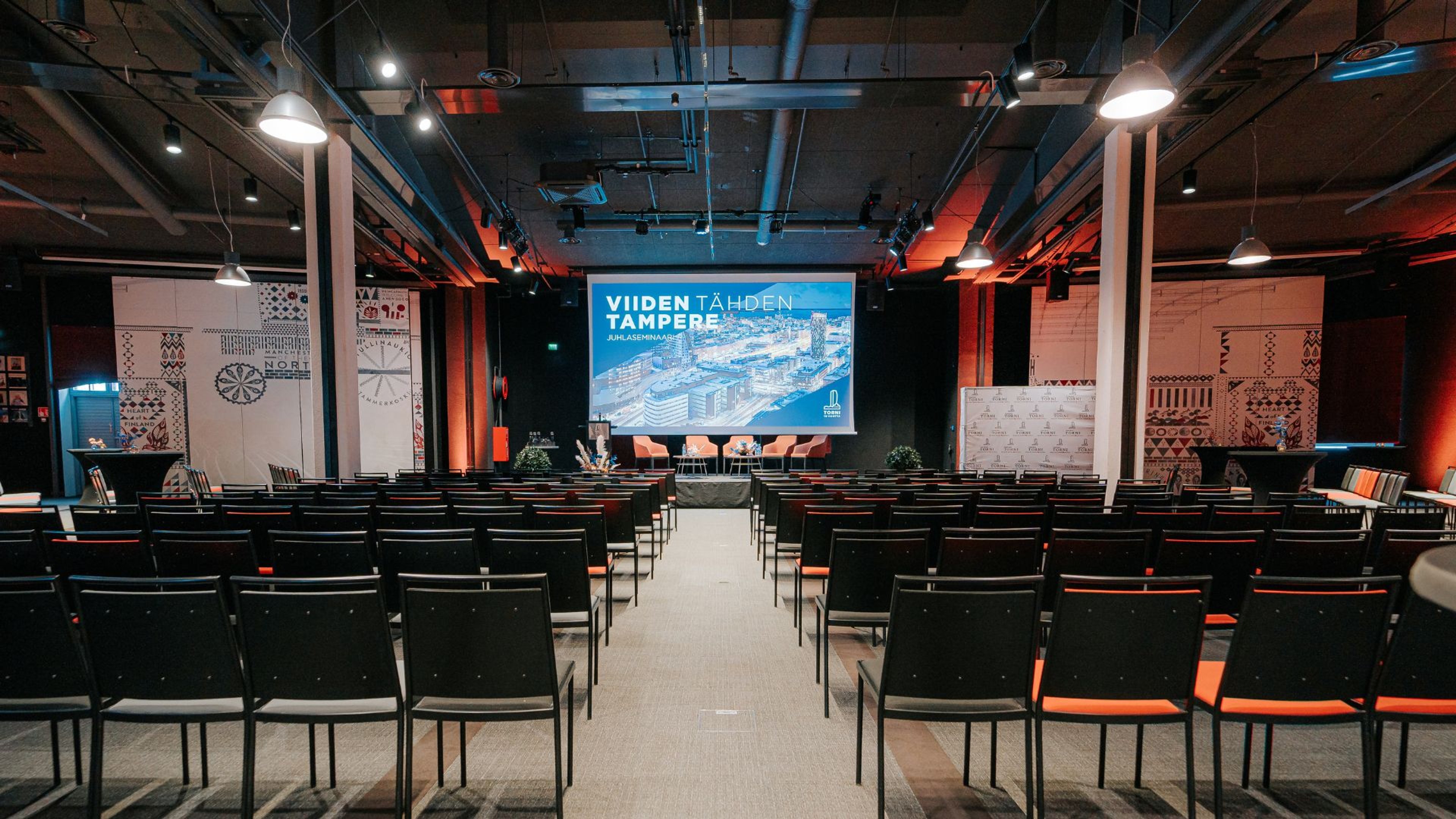 Moderni auditorio konferenssia varten, mustia tuoleja riveittäin, iso näyttö lavalla, teollisuushenkinen sisäkatto ja punasävyinen valaistus reunoilla.