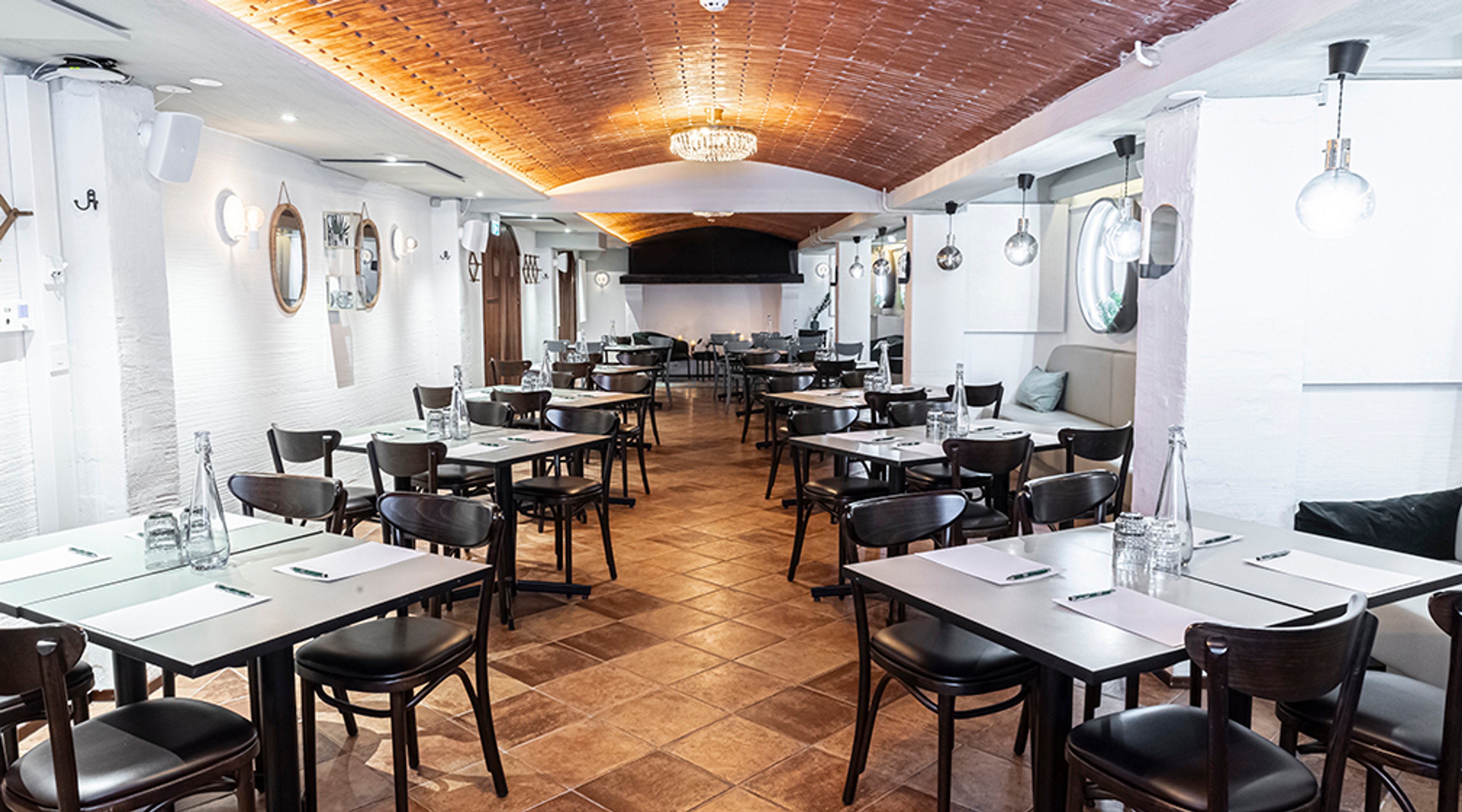 Ravintolasali, jossa valkoisia seiniä ja puinen kattopaneeli. Valaistus tulee kattokruunusta ja riippuvista lampuista. Tilassa on useita pöytiä mustilla tuoleilla.