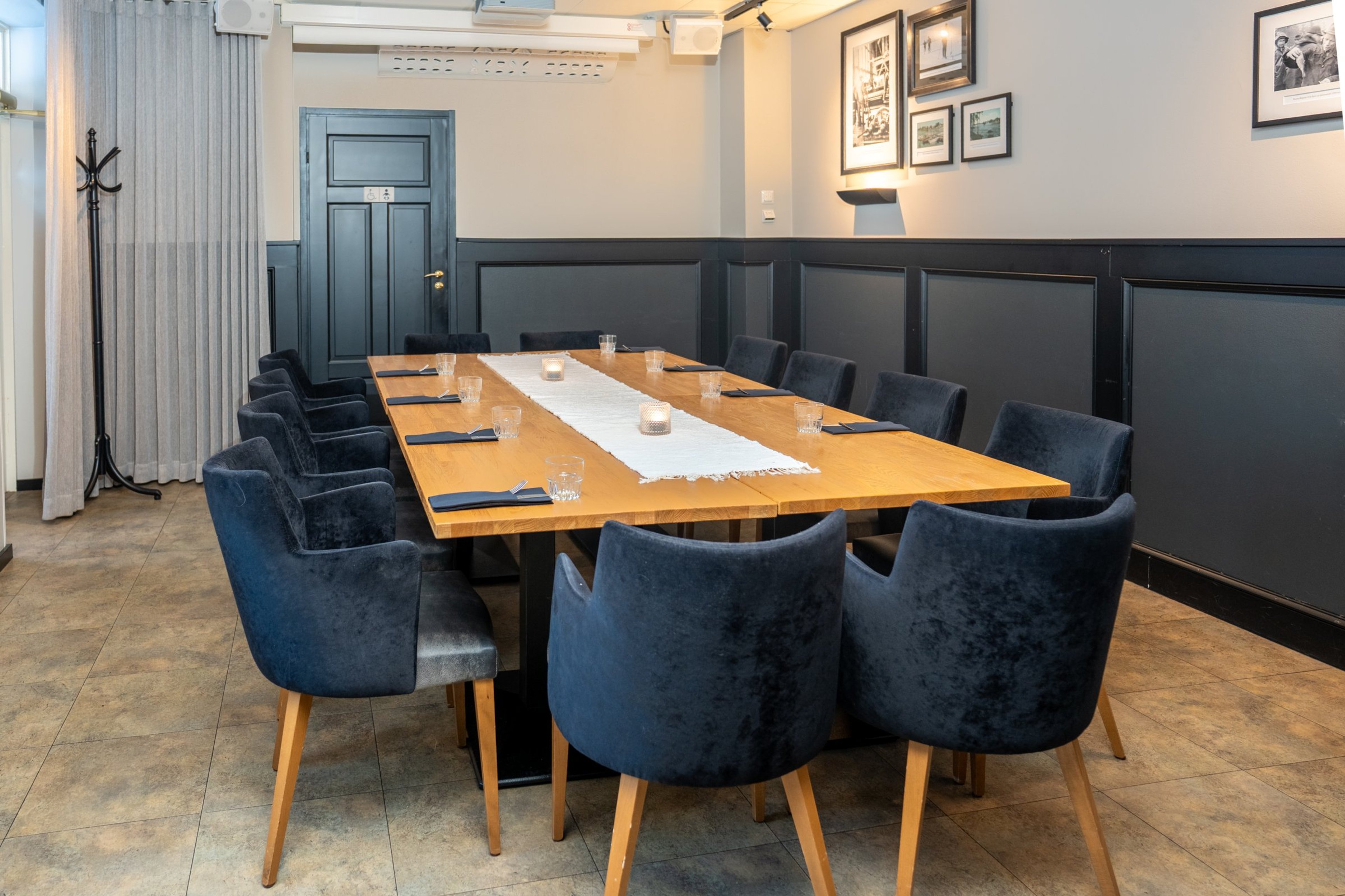 Kokoushuone jossa puinen pöytä ja 12 sinistä tuolia. Huoneessa on sininen ovi, tauluja seinällä ja kova lattiapinta, sekä inva-wc.