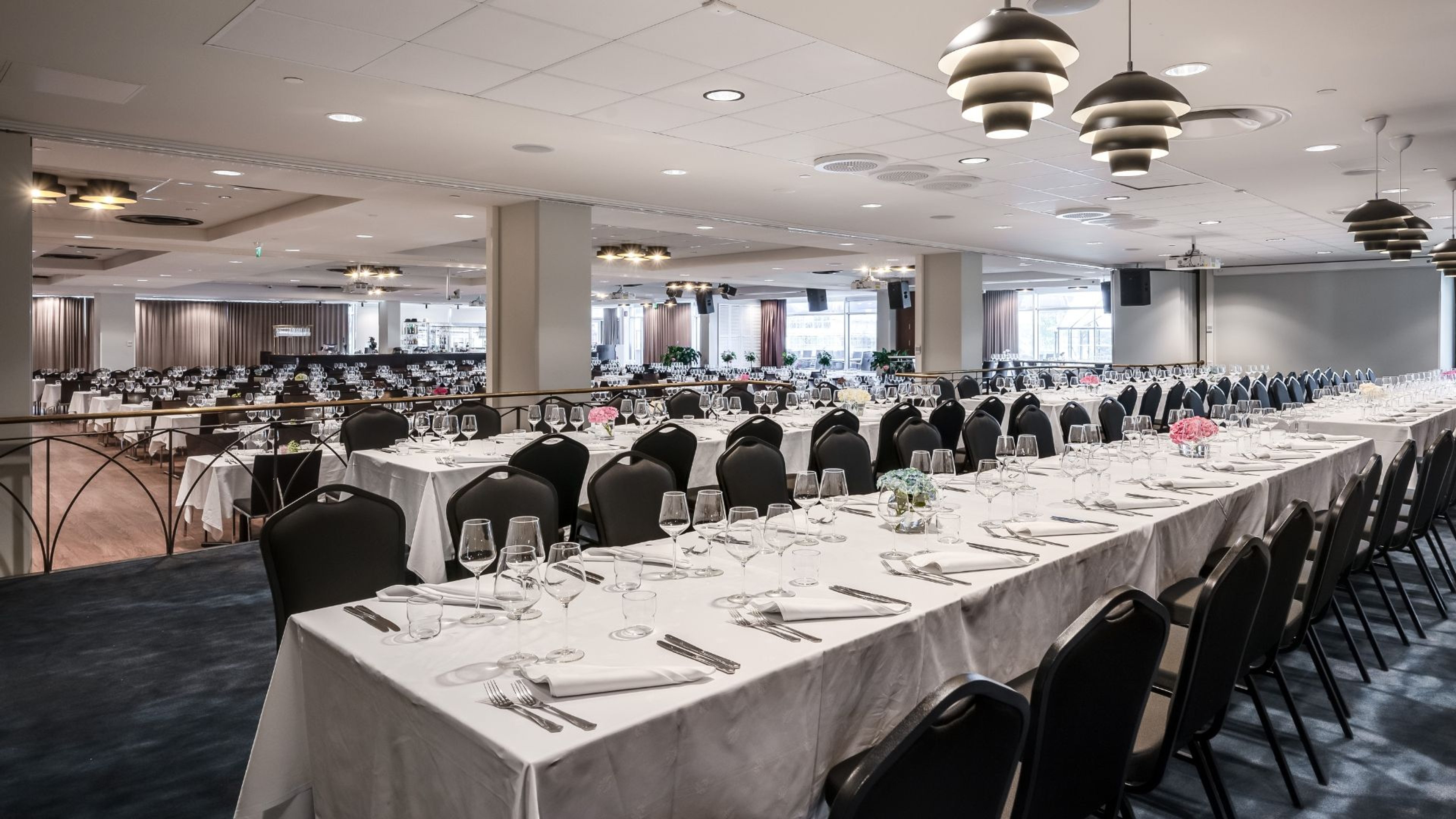 Tilava juhlatila valkoisin pöytäliinoin ja mustine tuoleineen. Pöydissä on kattaukset viinilaseineen ja kukkakoristein. Tila on hyvin valaistu, tasalattiainen.