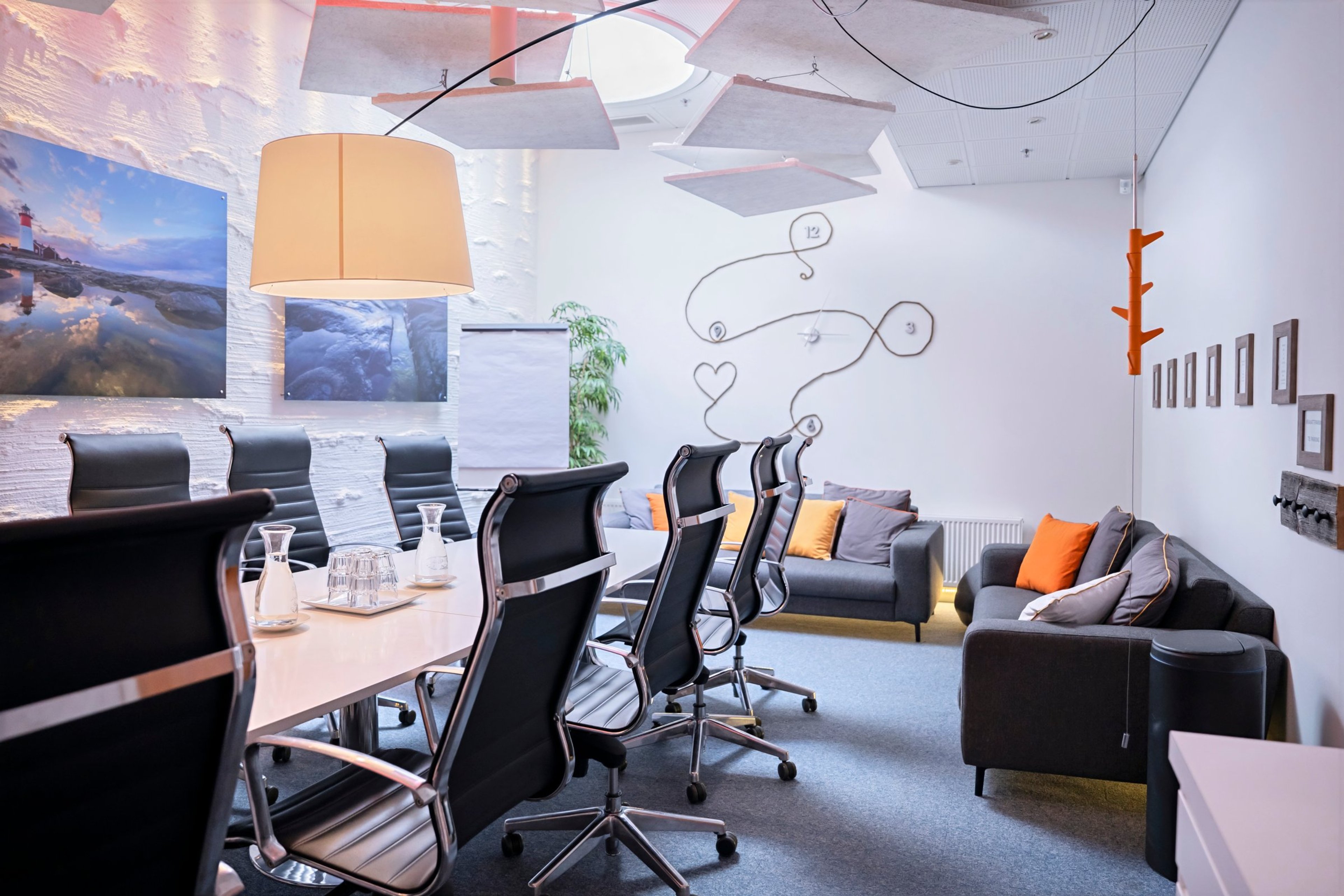 Moderni neuvotteluhuone, jossa on valkoinen pöytä, mustat toimistotuolit, harmaa sohvaryhmä oransseilla tyynyillä ja valkoinen sisustus taidevalokuvineen.