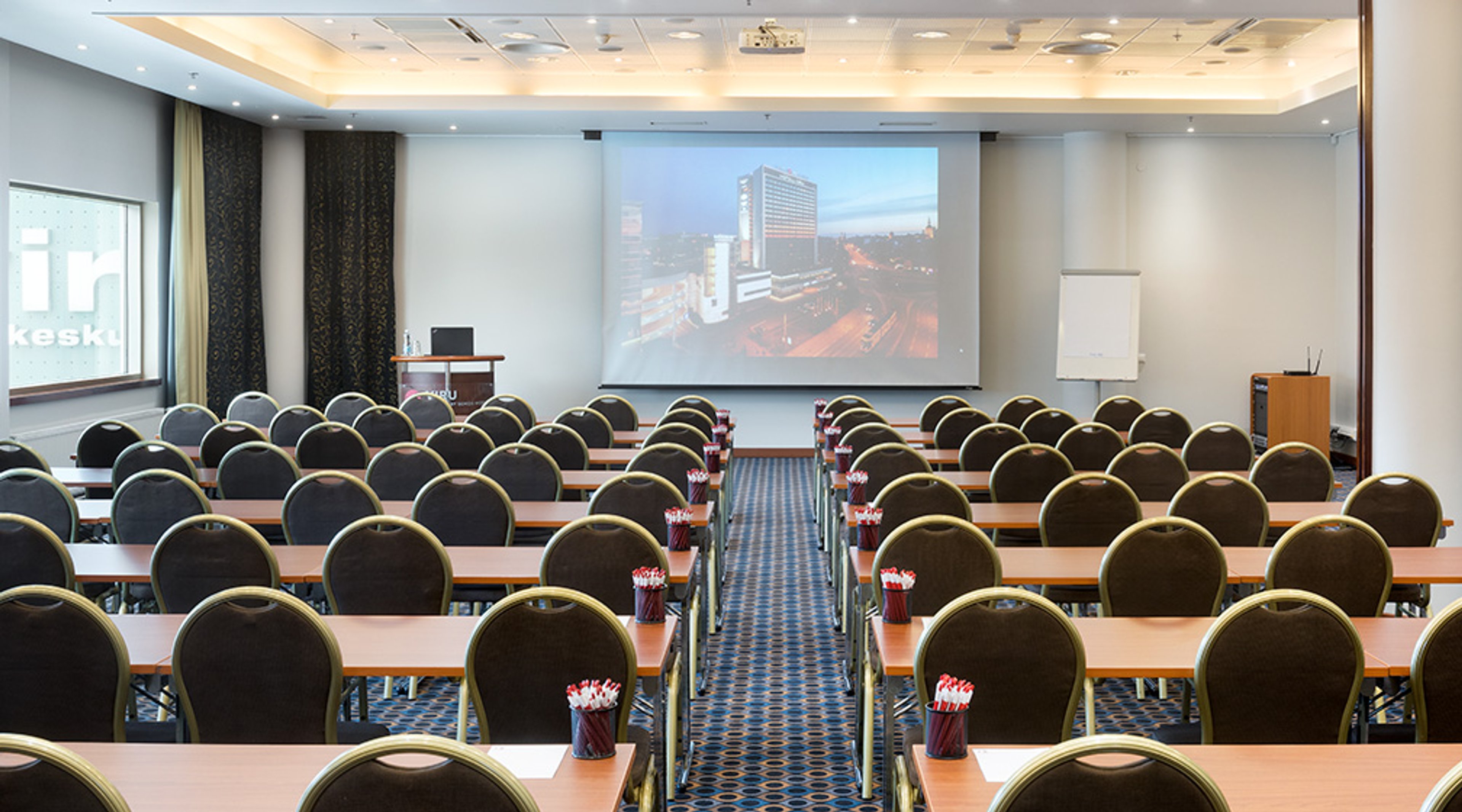 Auditorio kokousasetelmassa, jossa riveittäin pöytiä ja tuoleja. Edessä valkokangas, jossa kaupunkimaisema. Tila on hyvin valaistu ja lattialla sininen kokolattiamatto.