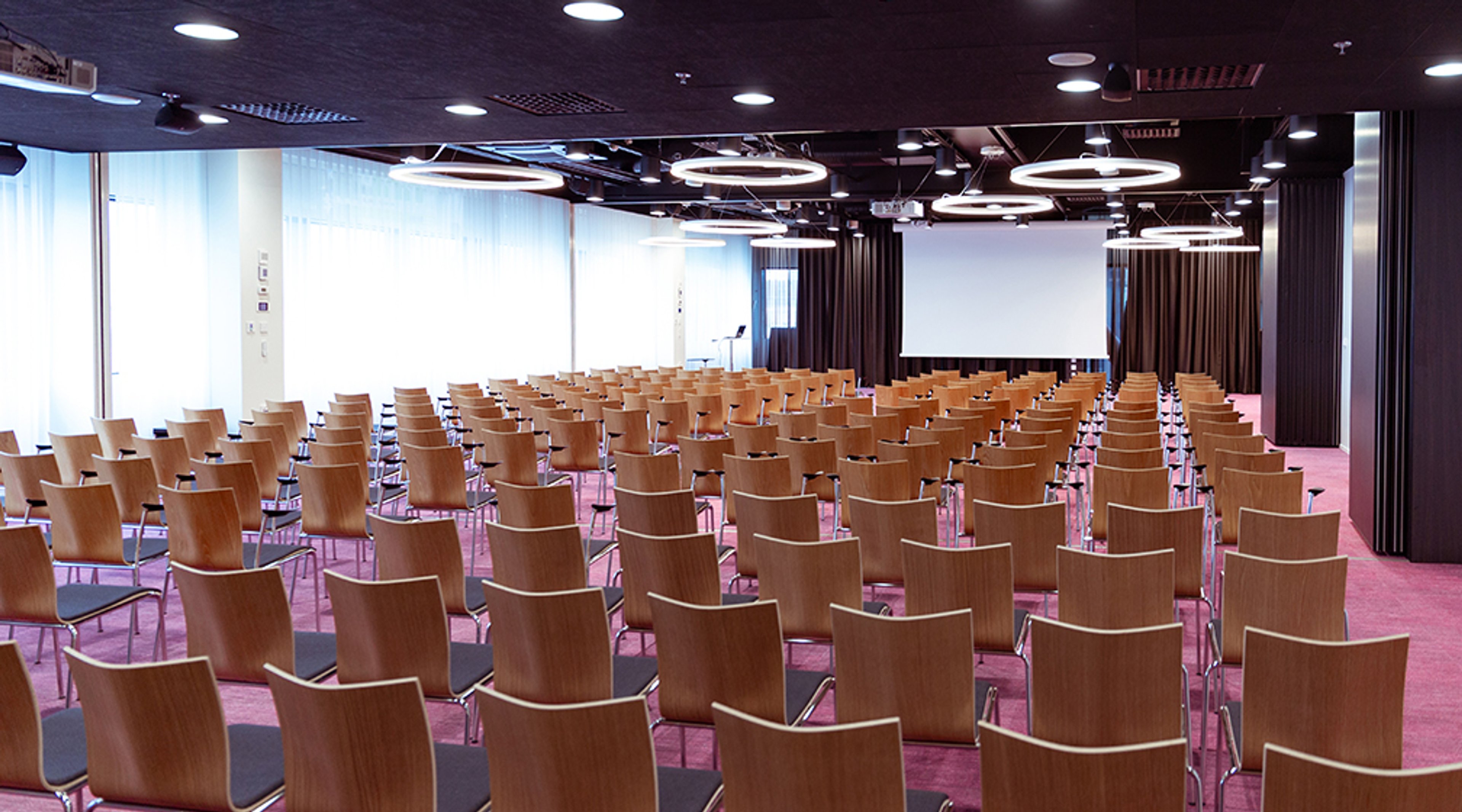 Large auditorium with wooden chairs, pink carpet, and a projector screen. Spacious room with large windows and circular ceiling lights.