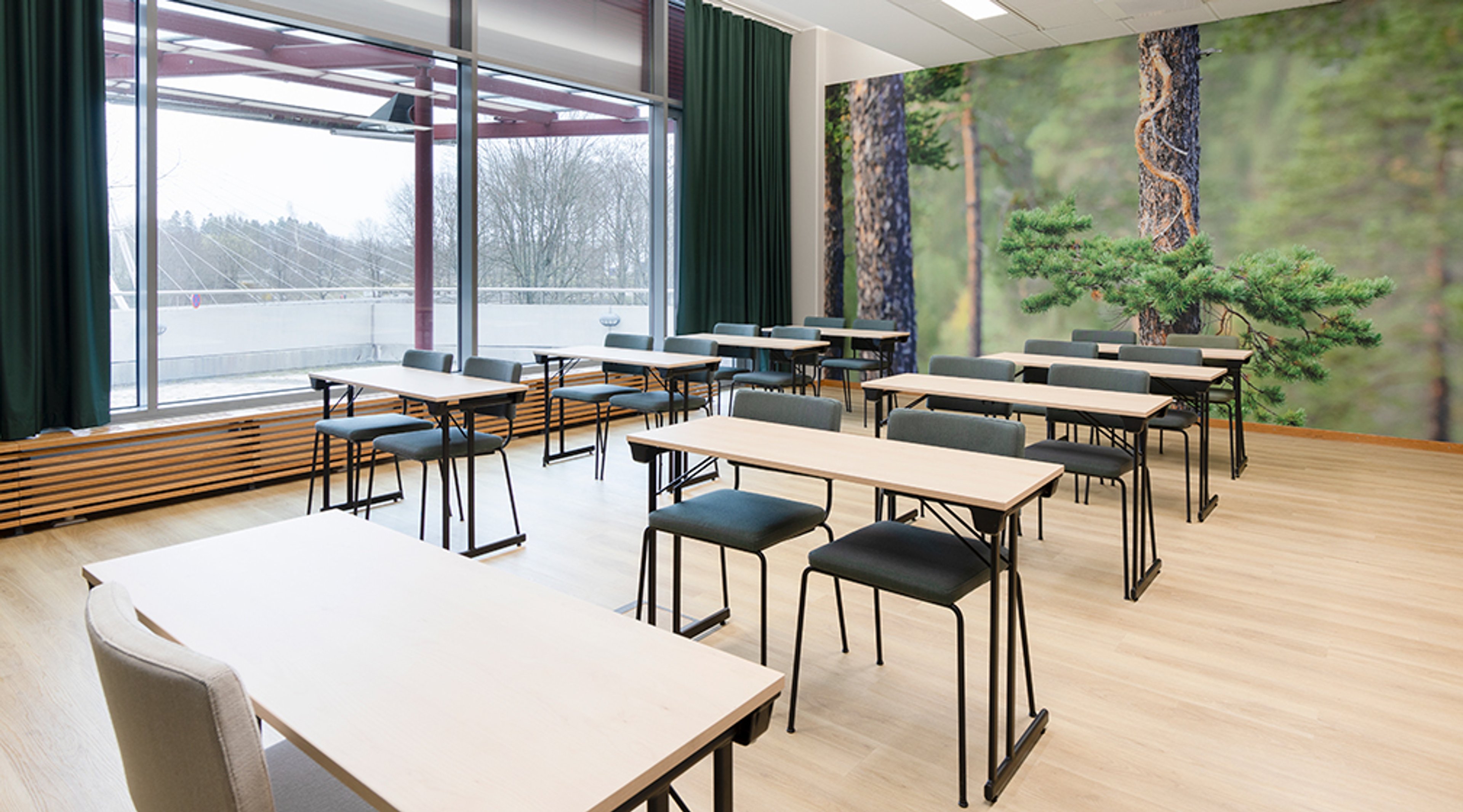 Valoisa kokoustila puisella lattialla, isoilla ikkunoilla ja metsäaiheisella seinäkuvalla. Tilassa on kevyet pöydät ja tuolit luokkamuodossa.
